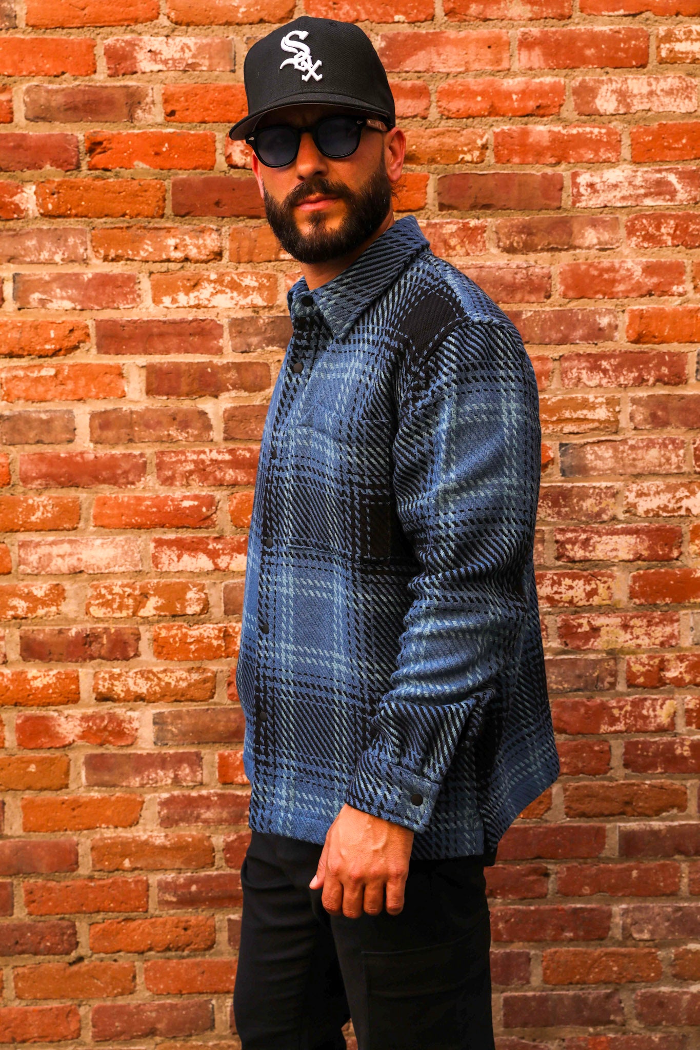 Man wearing a blue plaid shirt, black cap, and sunglasses against a brick wall.