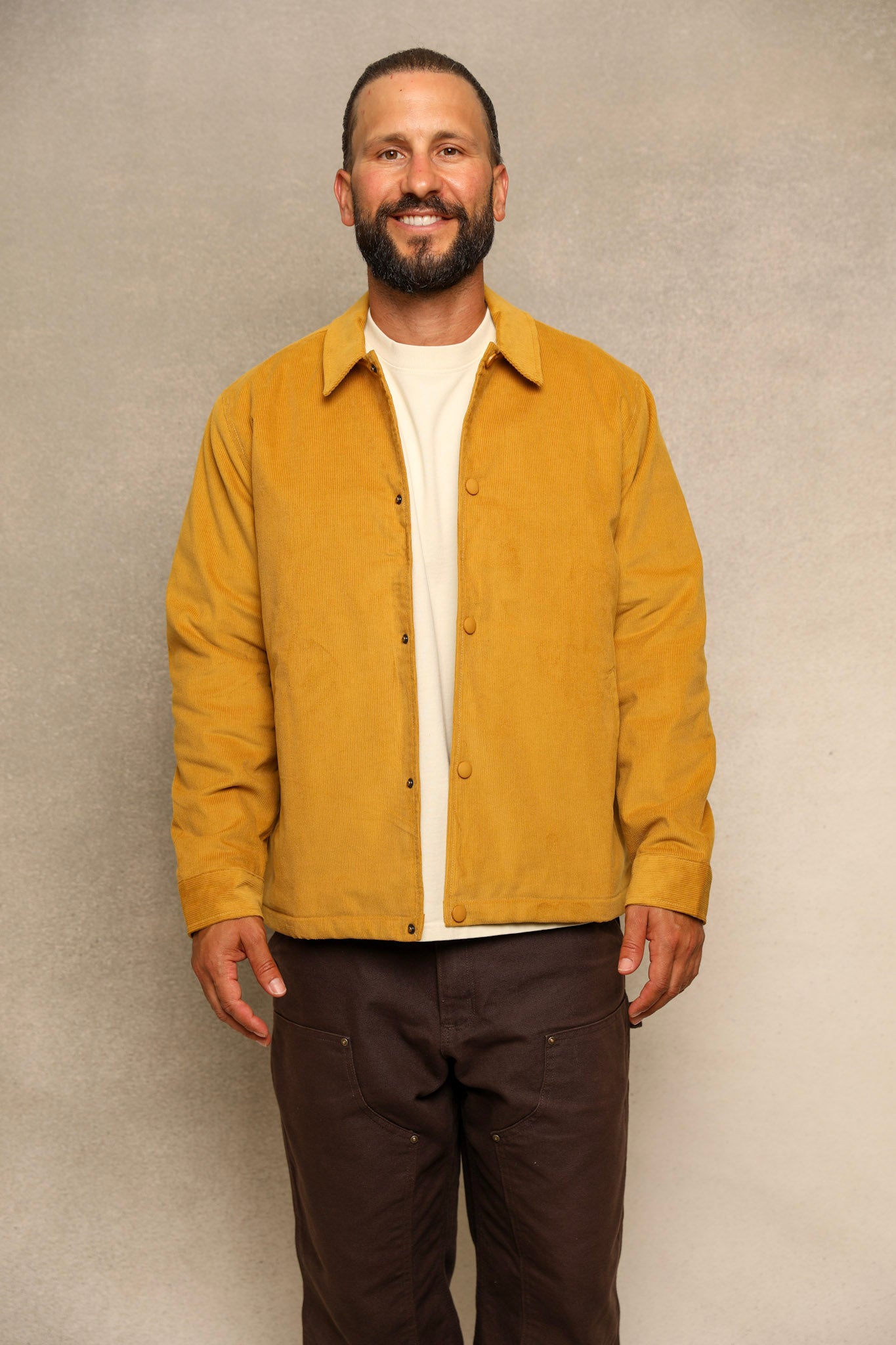 A smiling man wearing a mustard yellow button-up overshirt over a cream t-shirt and dark brown pants, standing against a textured gray studio background.