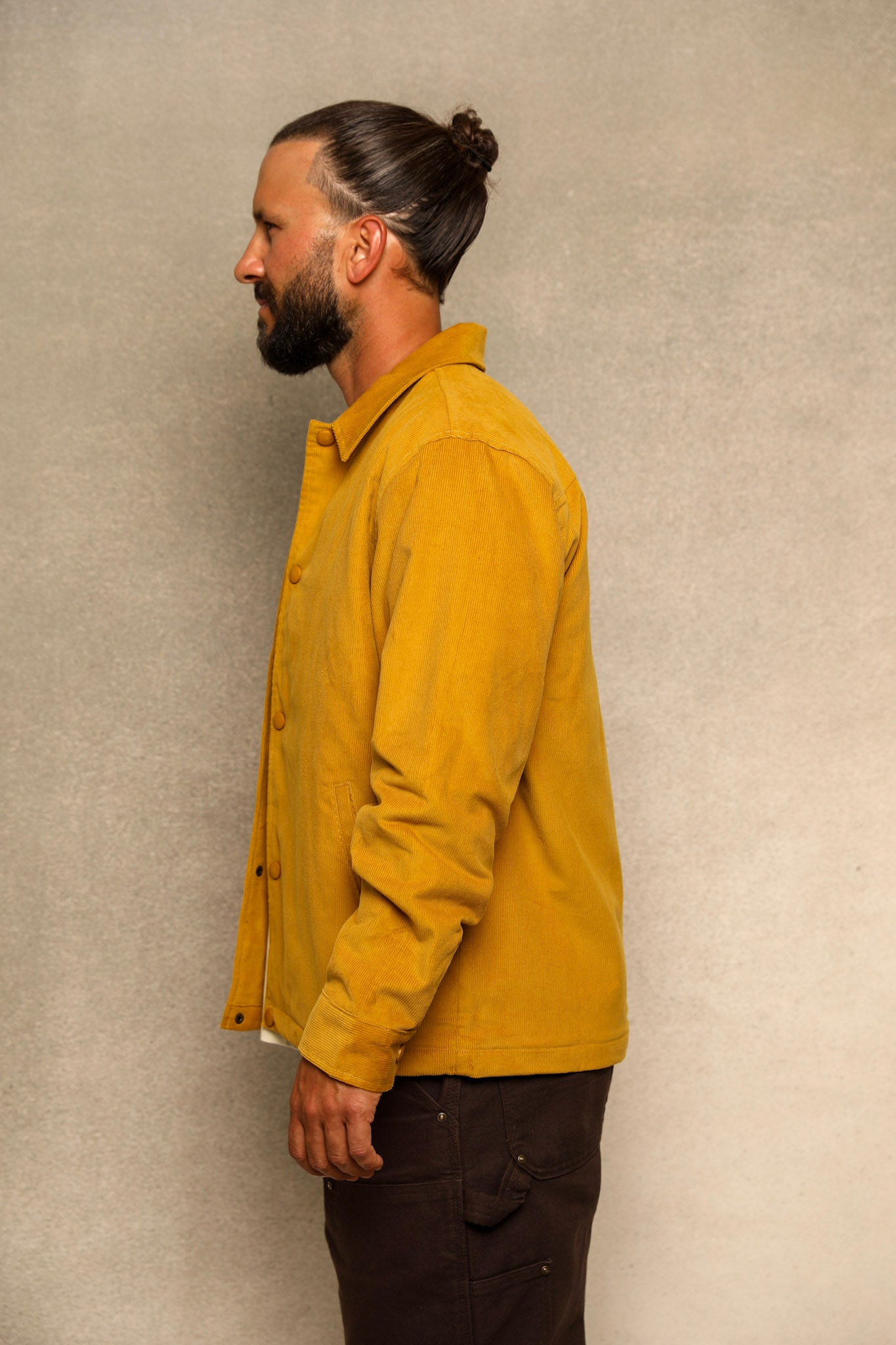 A man in profile wearing a mustard yellow button-up shirt and dark brown pants, standing against a textured light gray wall.