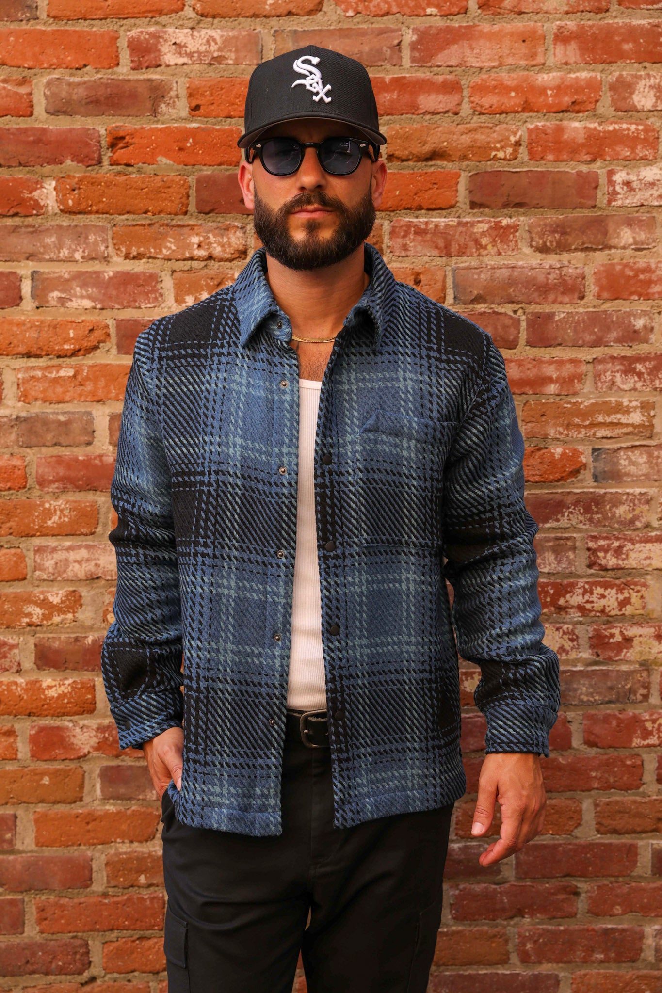 Man wearing a blue plaid shirt, black cap, and sunglasses against a brick wall.