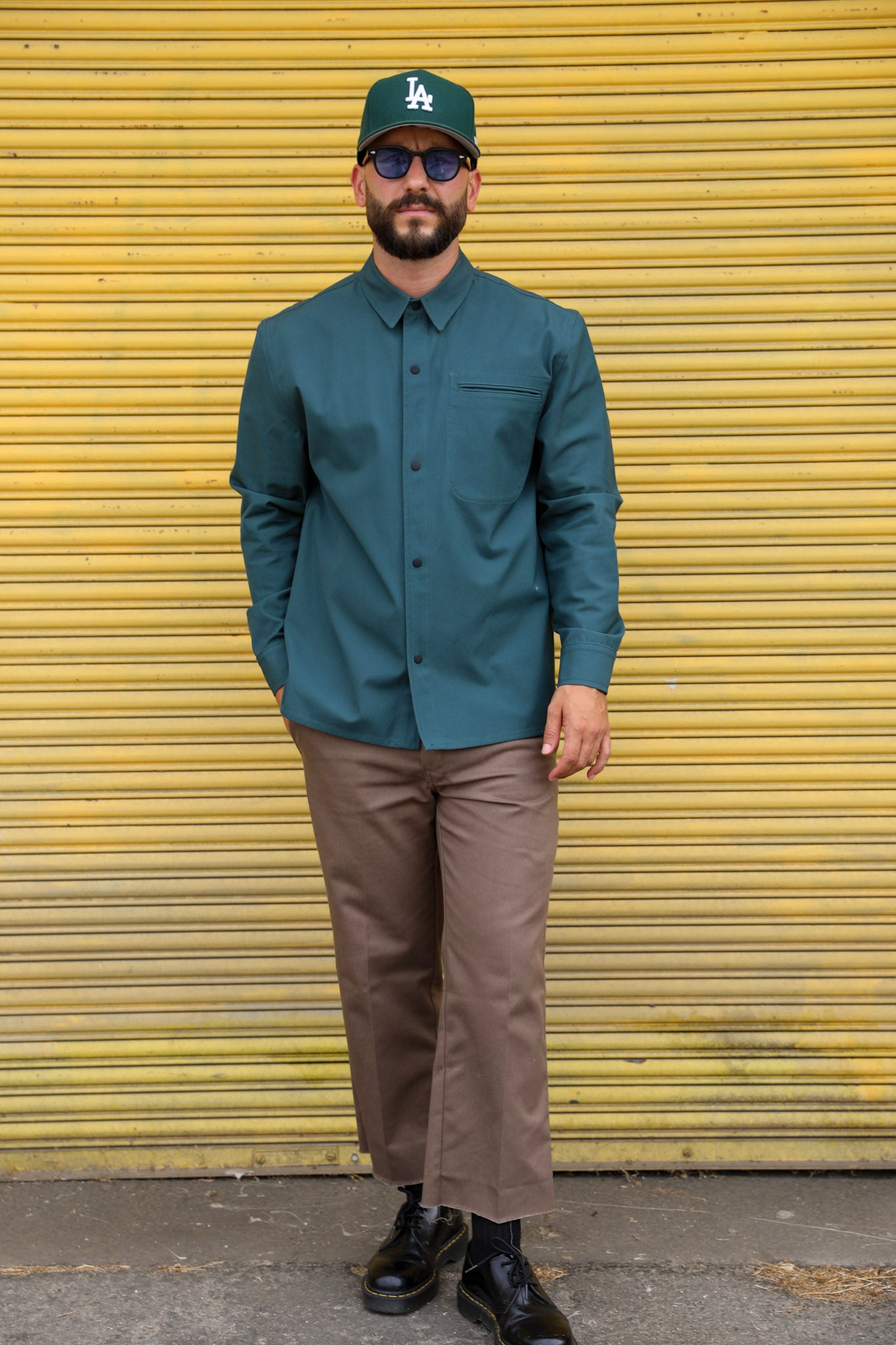 Man wearing a green shirt, brown pants, and a cap in front of a yellow shutter.