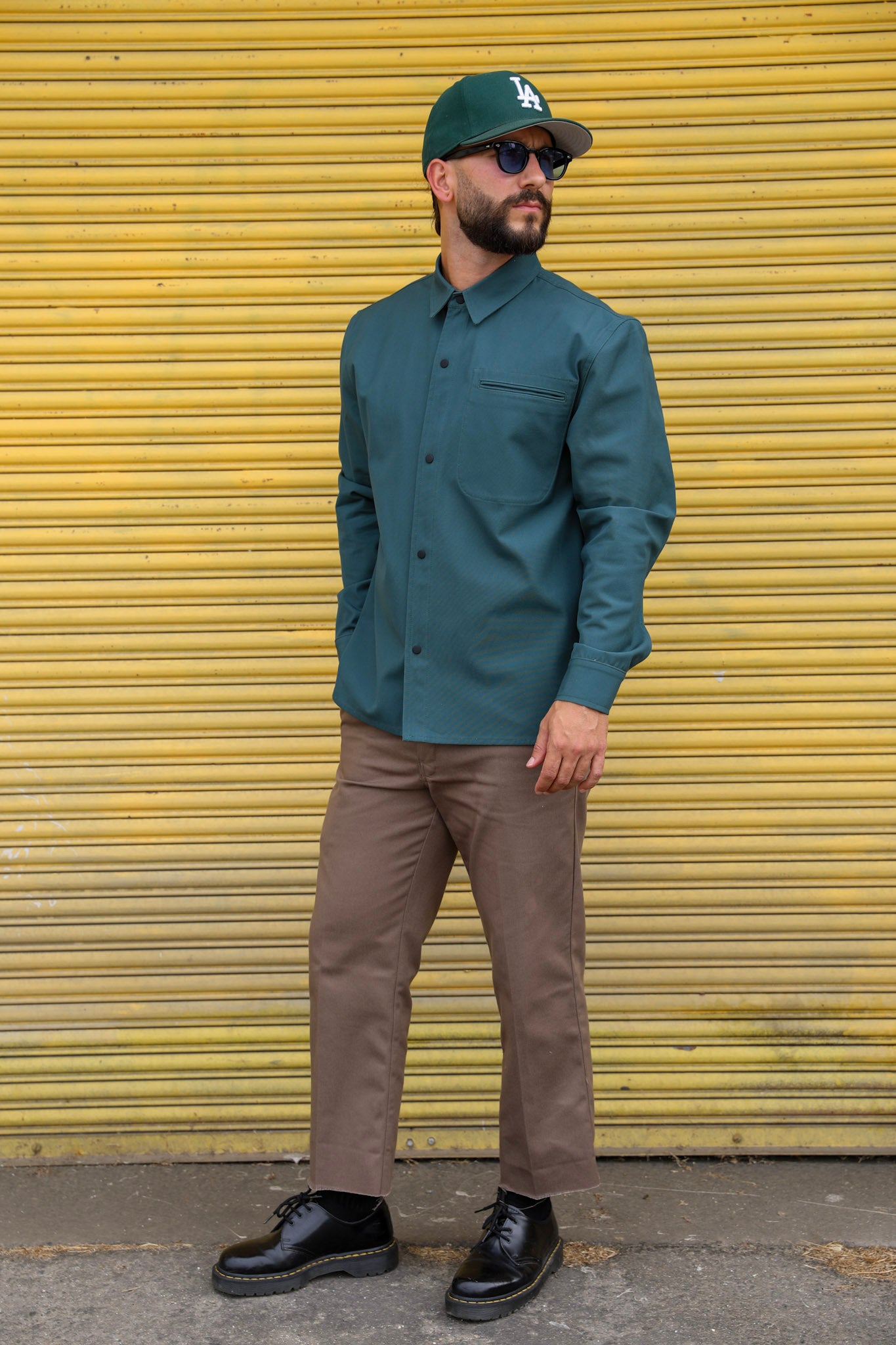 Man wearing a green shirt, brown pants, and a cap in front of a yellow shutter.