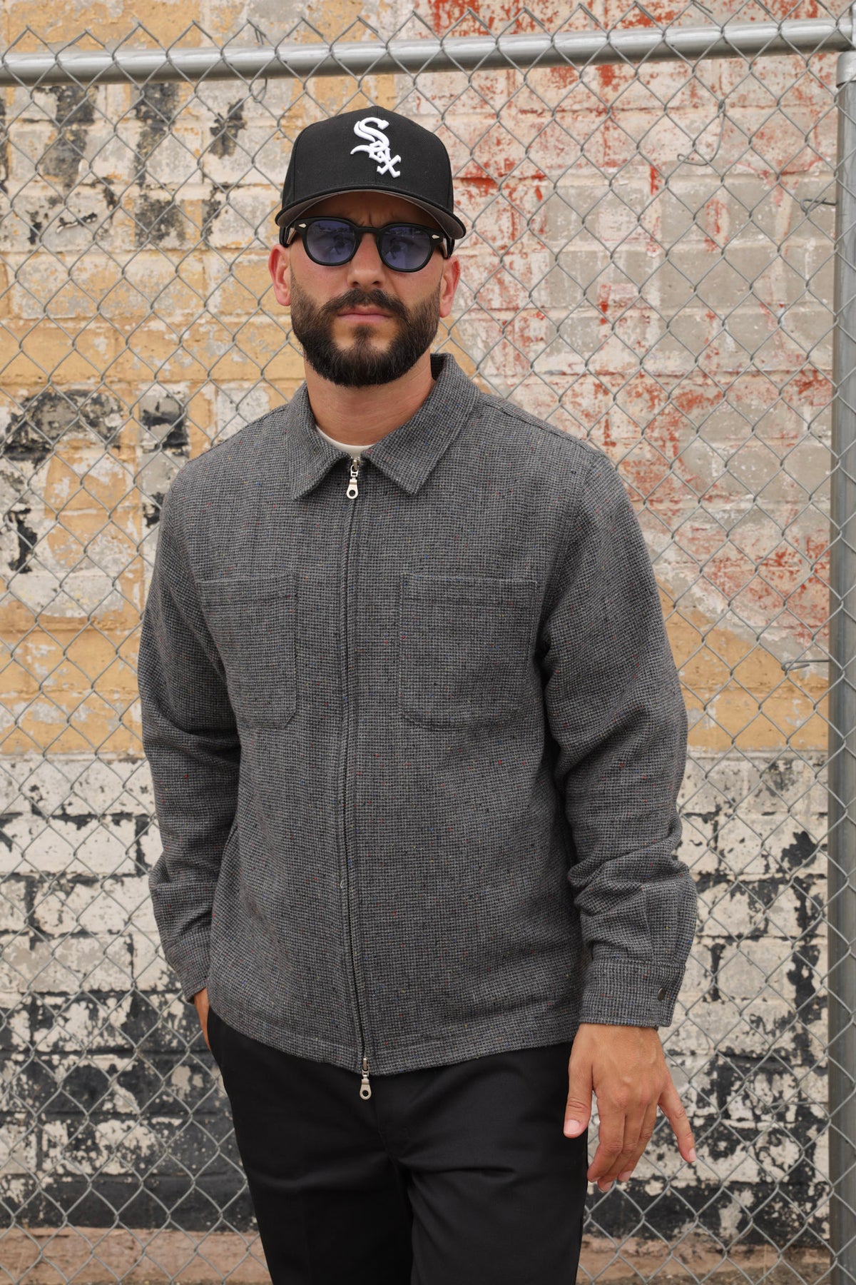 Man wearing a gray wool Signature zip-up jacket and black cap standing against a textured wall.