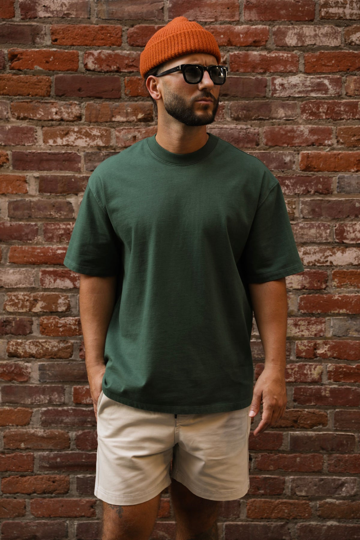 Man wearing a green t-shirt, beige shorts, orange beanie, and sunglasses against a brick wall.
