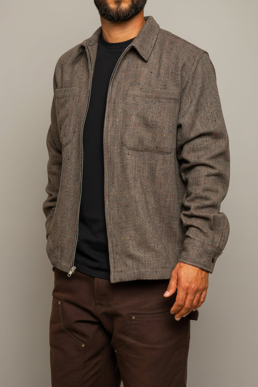 A man with a beard wearing a textured gray overshirt over a black t-shirt and dark brown pants, standing against a neutral textured gray wall.