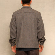 A man viewed from behind wearing a textured gray long-sleeve overshirt and dark brown cargo pants, standing against a neutral textured beige wall.