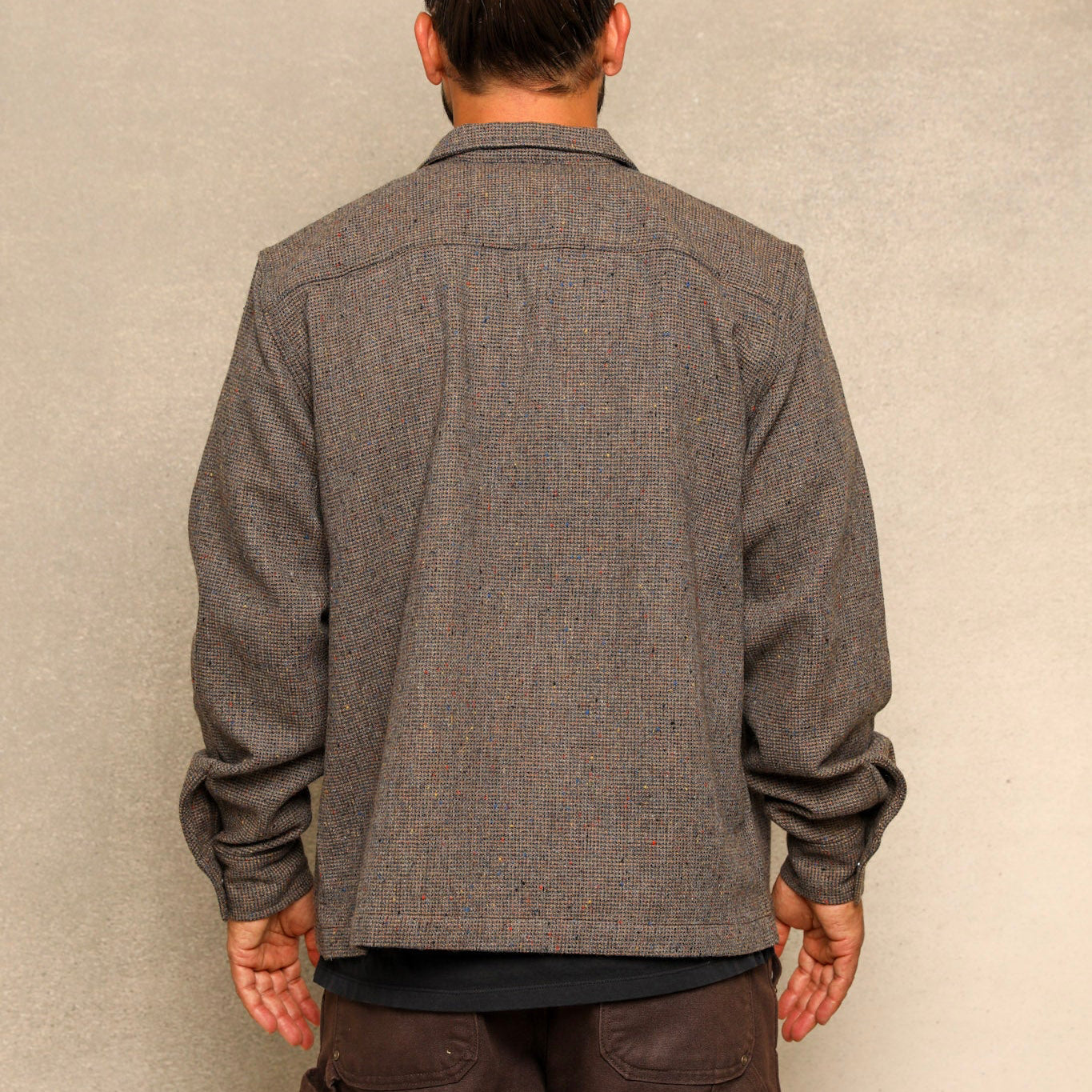 A man viewed from behind wearing a textured gray long-sleeve overshirt and dark brown cargo pants, standing against a neutral textured beige wall.