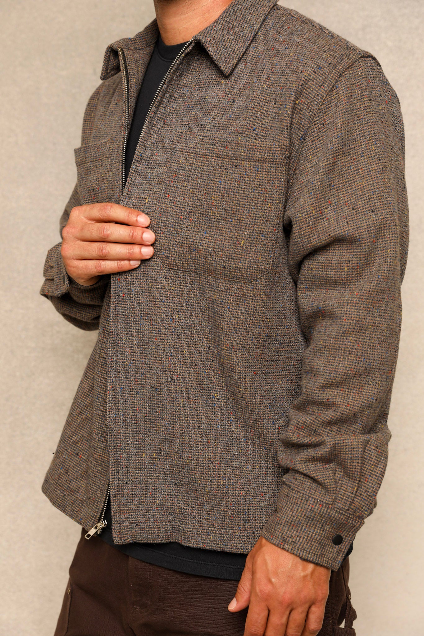 A man wearing a brown and gray textured zip-up overshirt jacket over a black shirt, shown from the neck down against a neutral gray background.