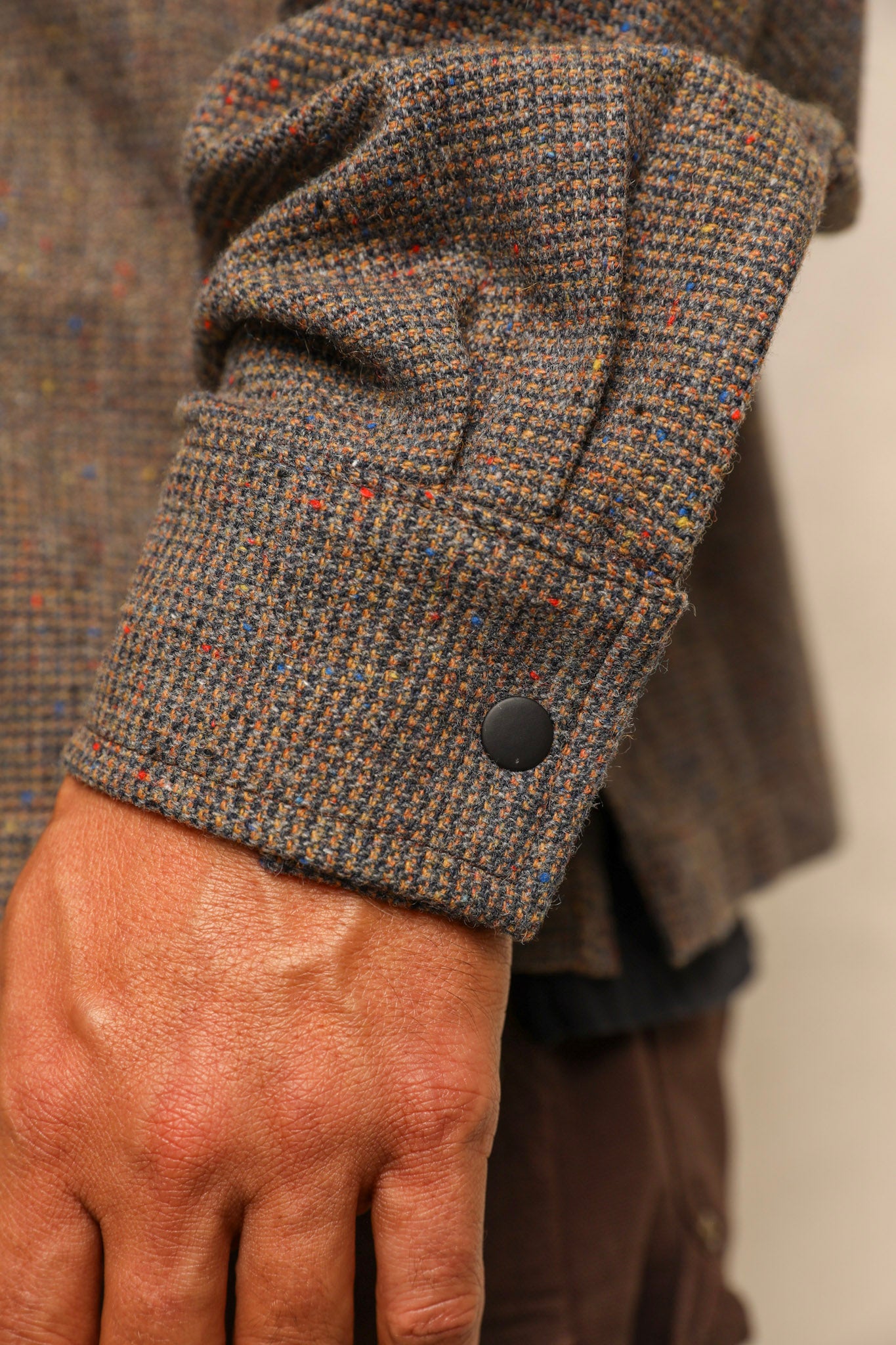 Close-up of a person's hand wearing a brown tweed jacket with a rolled cuff showing a black button closure.
