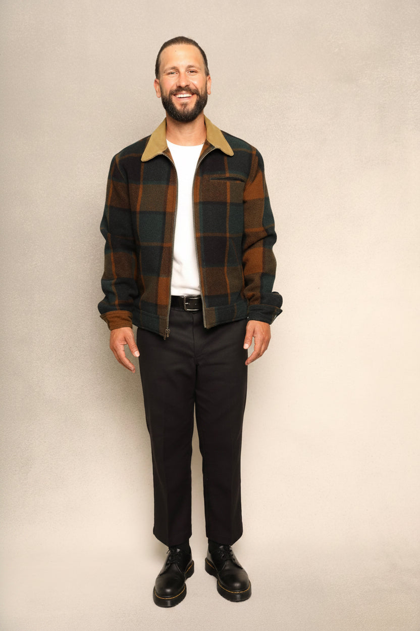 A man wearing a brown and black plaid overshirt with a tan collar, a white t-shirt, black trousers, and black leather shoes, standing against a textured beige studio backdrop.