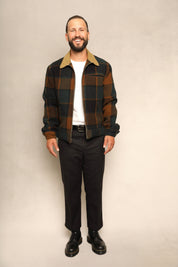 A man wearing a brown and black plaid overshirt with a tan collar, a white t-shirt, black trousers, and black leather shoes, standing against a textured beige studio backdrop.