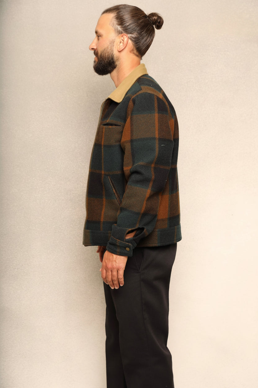 A man in profile wearing a dark green and brown plaid jacket with a tan collar, paired with black trousers, standing against a textured beige wall.