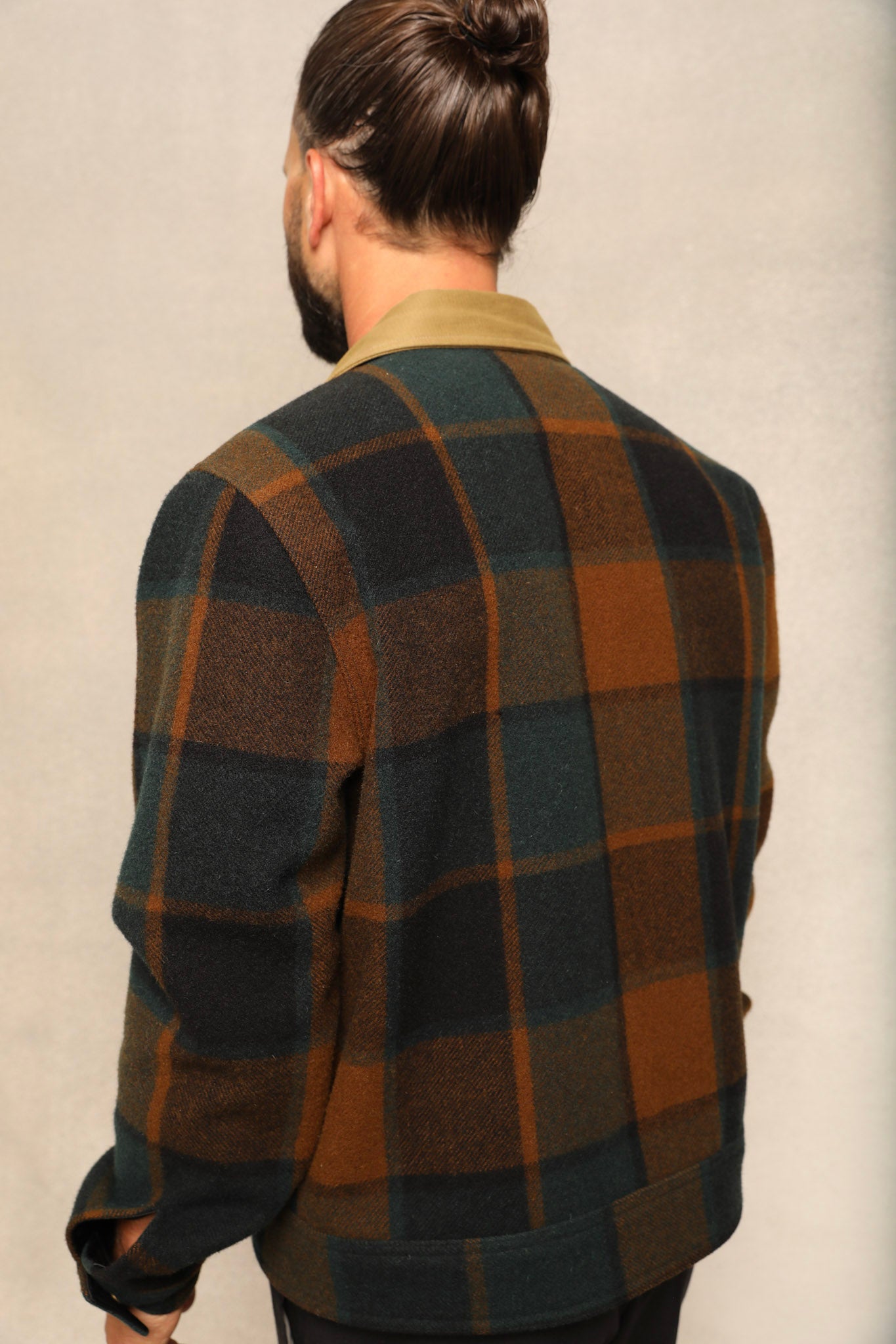 A man viewed from behind wearing a dark green and brown plaid wool-blend jacket over a tan collared shirt, set against a neutral textured studio background.