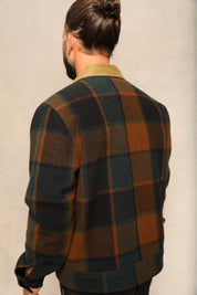 A man viewed from behind wearing a dark green and brown plaid wool-blend jacket over a tan collared shirt, set against a neutral textured studio background.