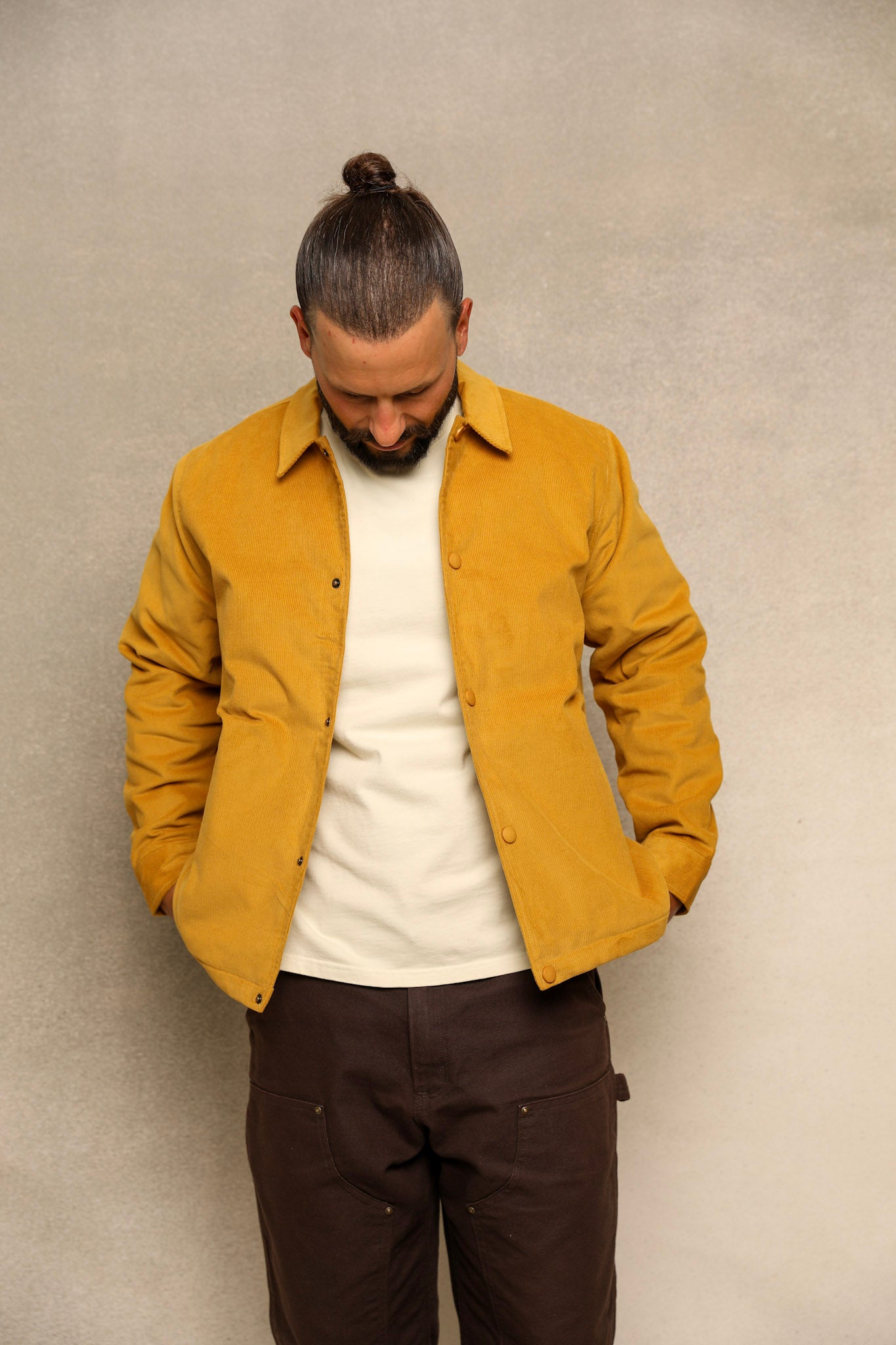 A man wearing a mustard yellow overshirt over a white t-shirt and dark brown pants, standing against a textured gray wall.