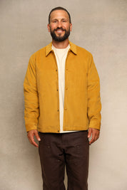 A smiling man wearing a mustard yellow button-up overshirt over a cream t-shirt and dark brown pants, standing against a textured gray studio background.