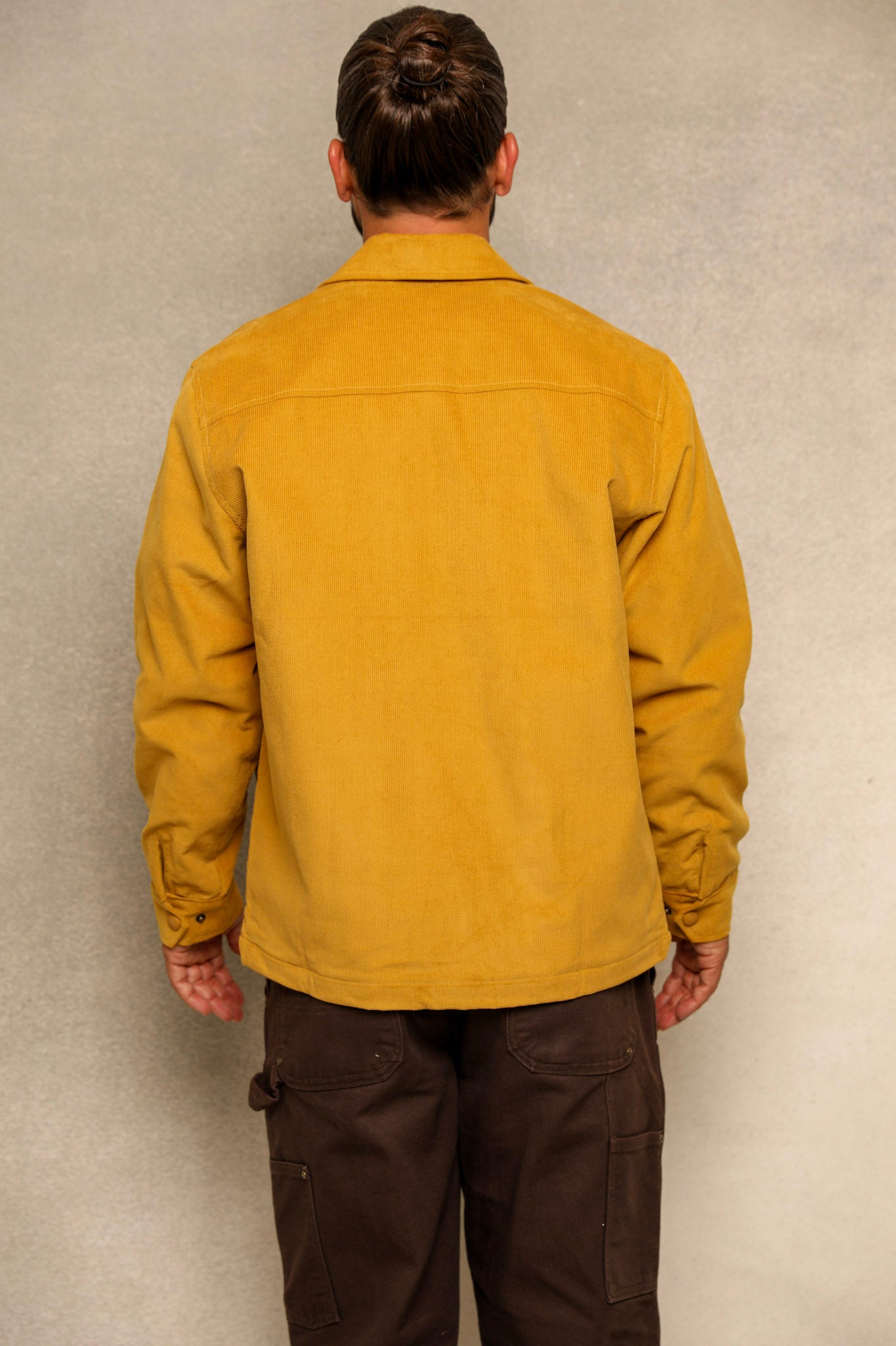 A man viewed from behind wearing a mustard yellow utility jacket and dark brown cargo pants, standing against a textured light gray wall.