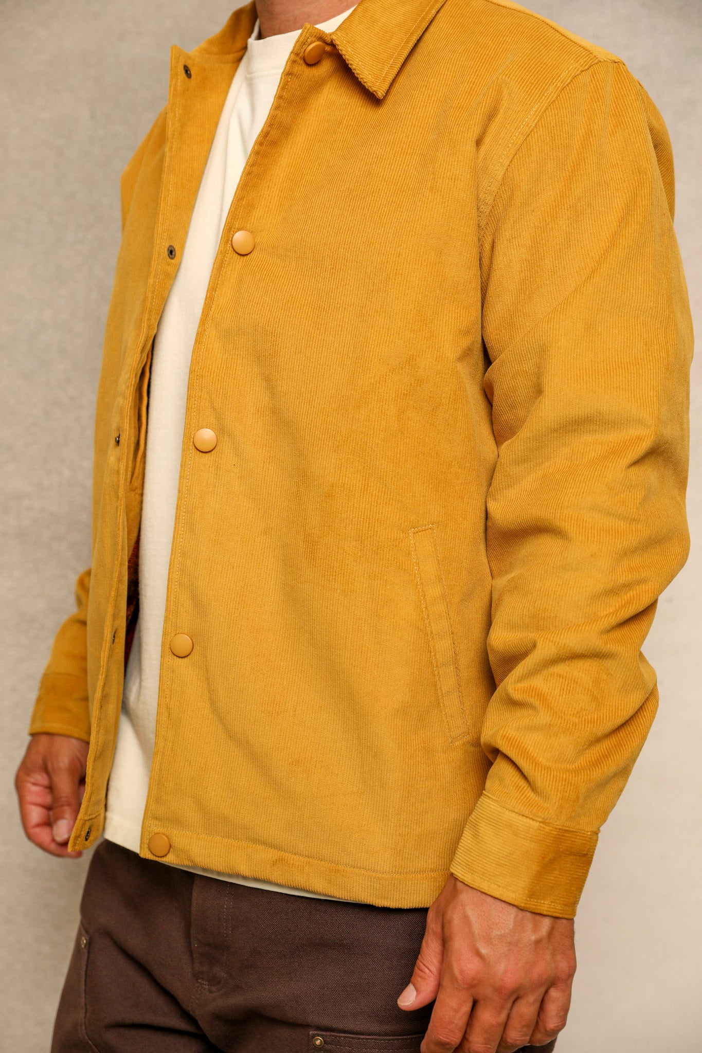A close-up of a person wearing a mustard yellow corduroy jacket over a white t-shirt and dark brown pants, set against a neutral gray background.