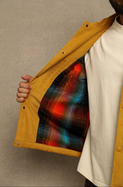 A close-up of a person wearing a mustard yellow overshirt over a white t-shirt, opening the overshirt to show a colorful plaid lining with red, blue, and green tones.