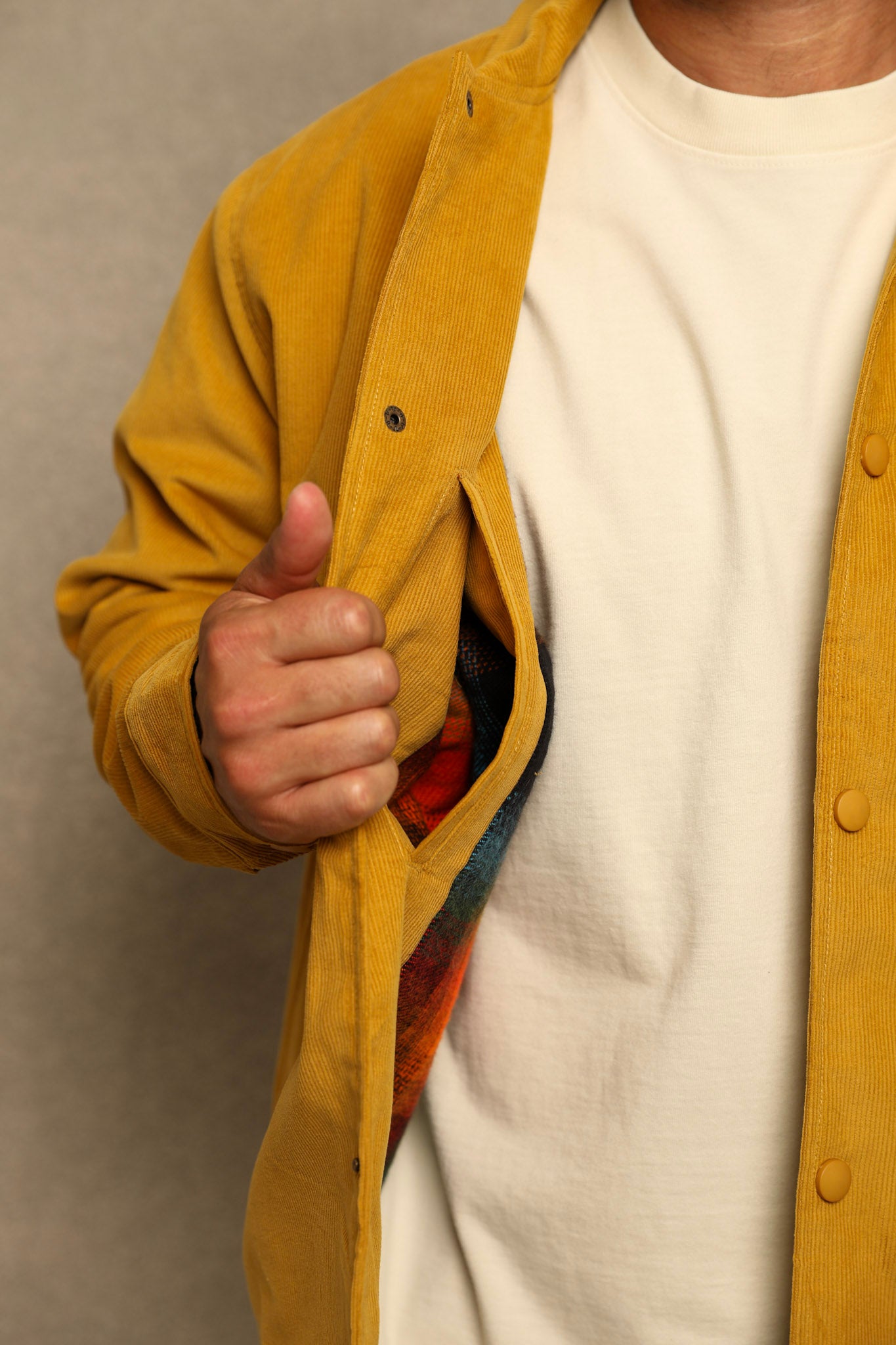 A close-up of a person wearing a mustard yellow corduroy overshirt over a cream t-shirt, with their hand pulling the overshirt open to reveal a colorful patterned lining.