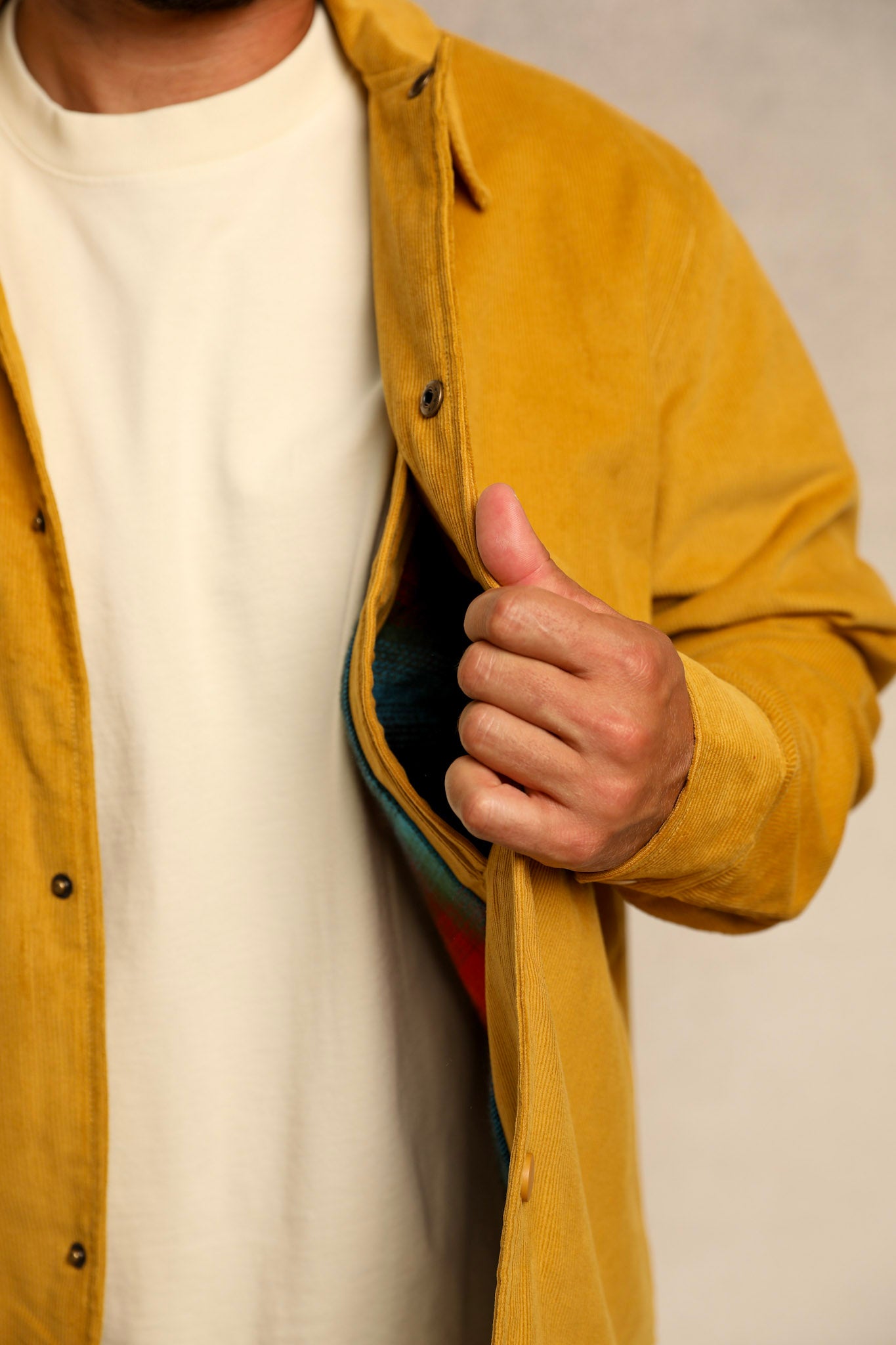 A close-up of a person wearing a mustard yellow overshirt over a white t-shirt, pulling the overshirt open to reveal a colorful striped shirt underneath.