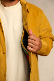 A close-up of a person wearing a mustard yellow overshirt over a white t-shirt, pulling the overshirt open to reveal a colorful striped shirt underneath.