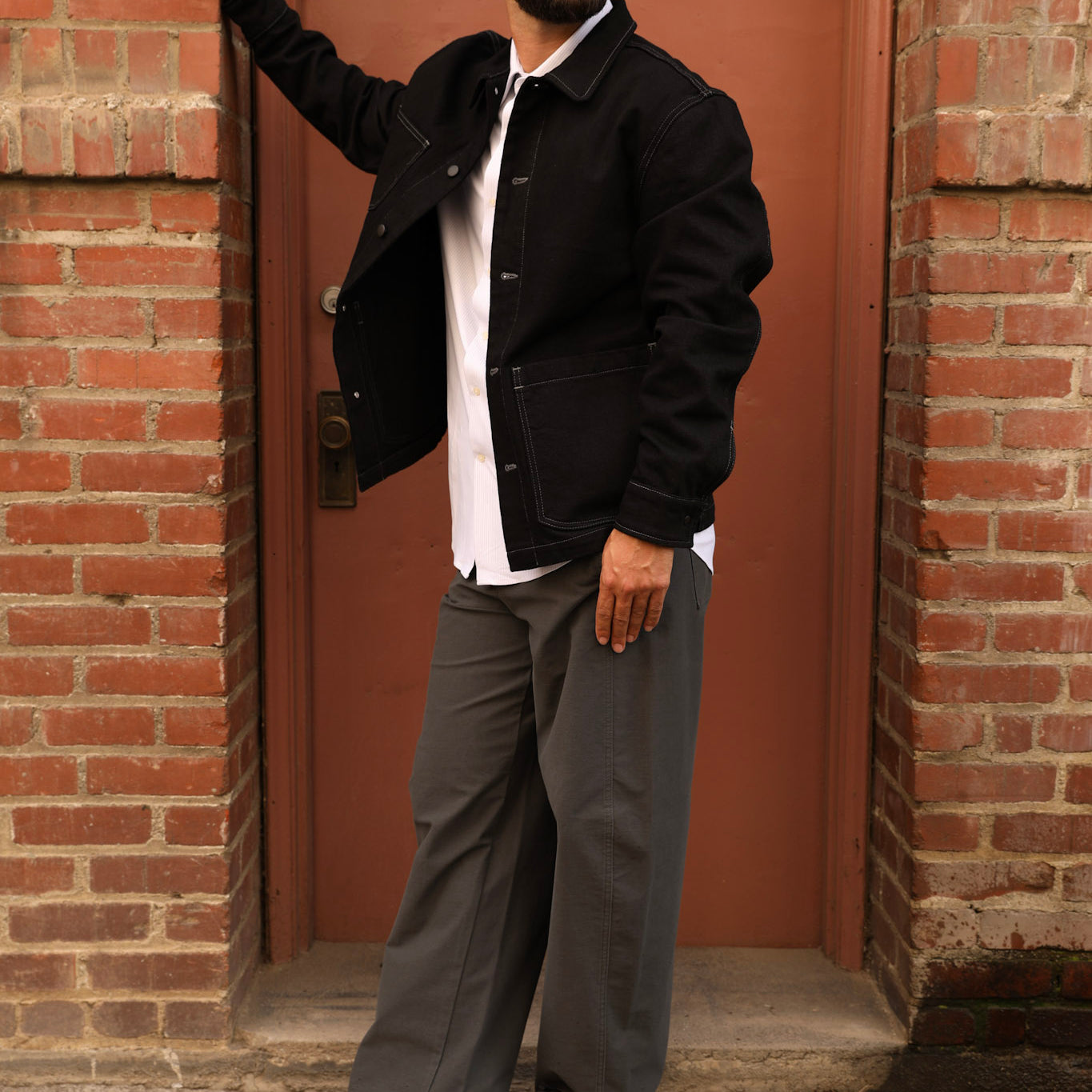 A man wearing a black baseball cap with a white 'A' logo, sunglasses, a black jacket over a white shirt, gray wide-leg pants, and black loafers, posing in front of a red brick door.