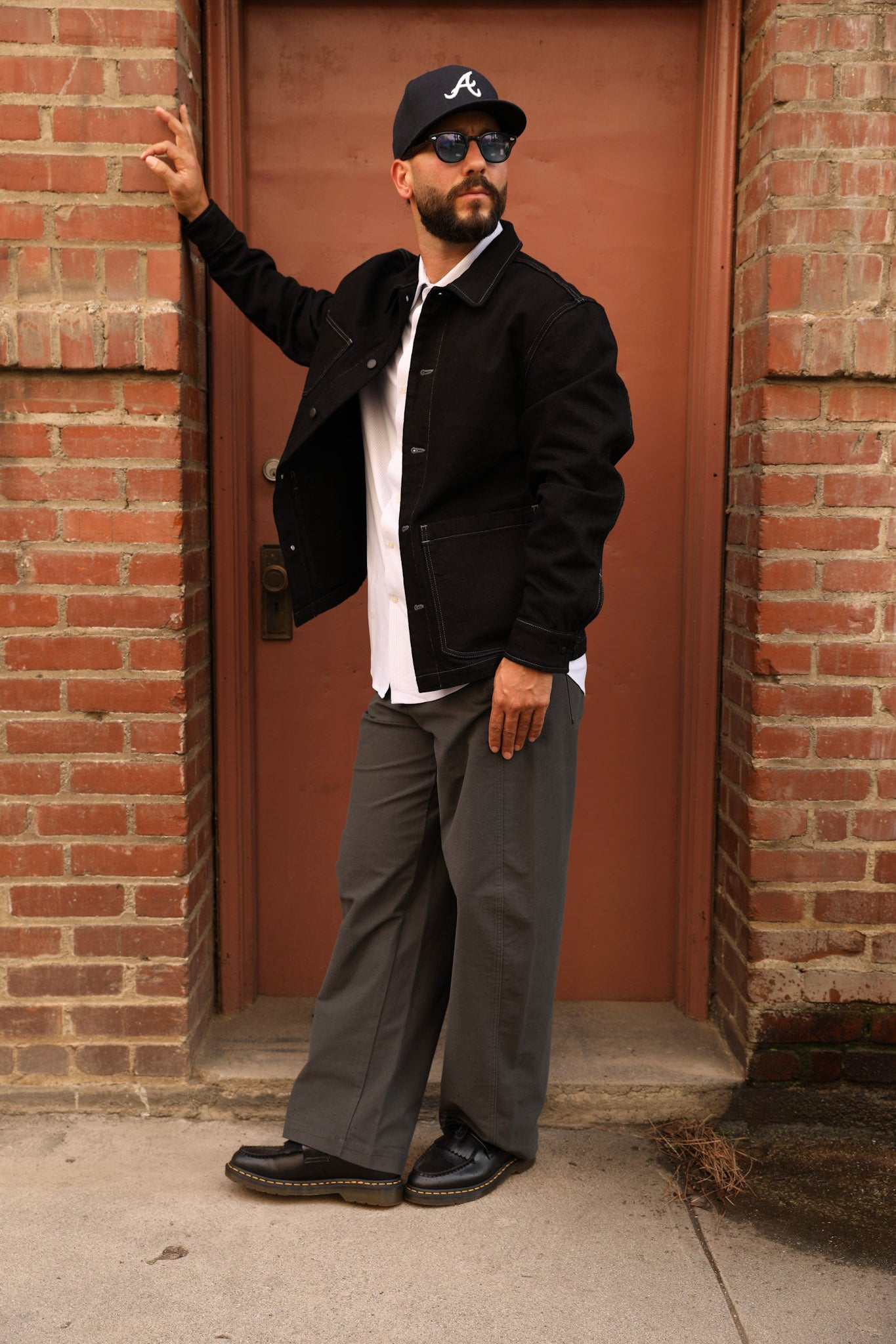 A man wearing a black baseball cap with a white 'A' logo, sunglasses, a black jacket over a white shirt, gray wide-leg pants, and black loafers, posing in front of a red brick door.