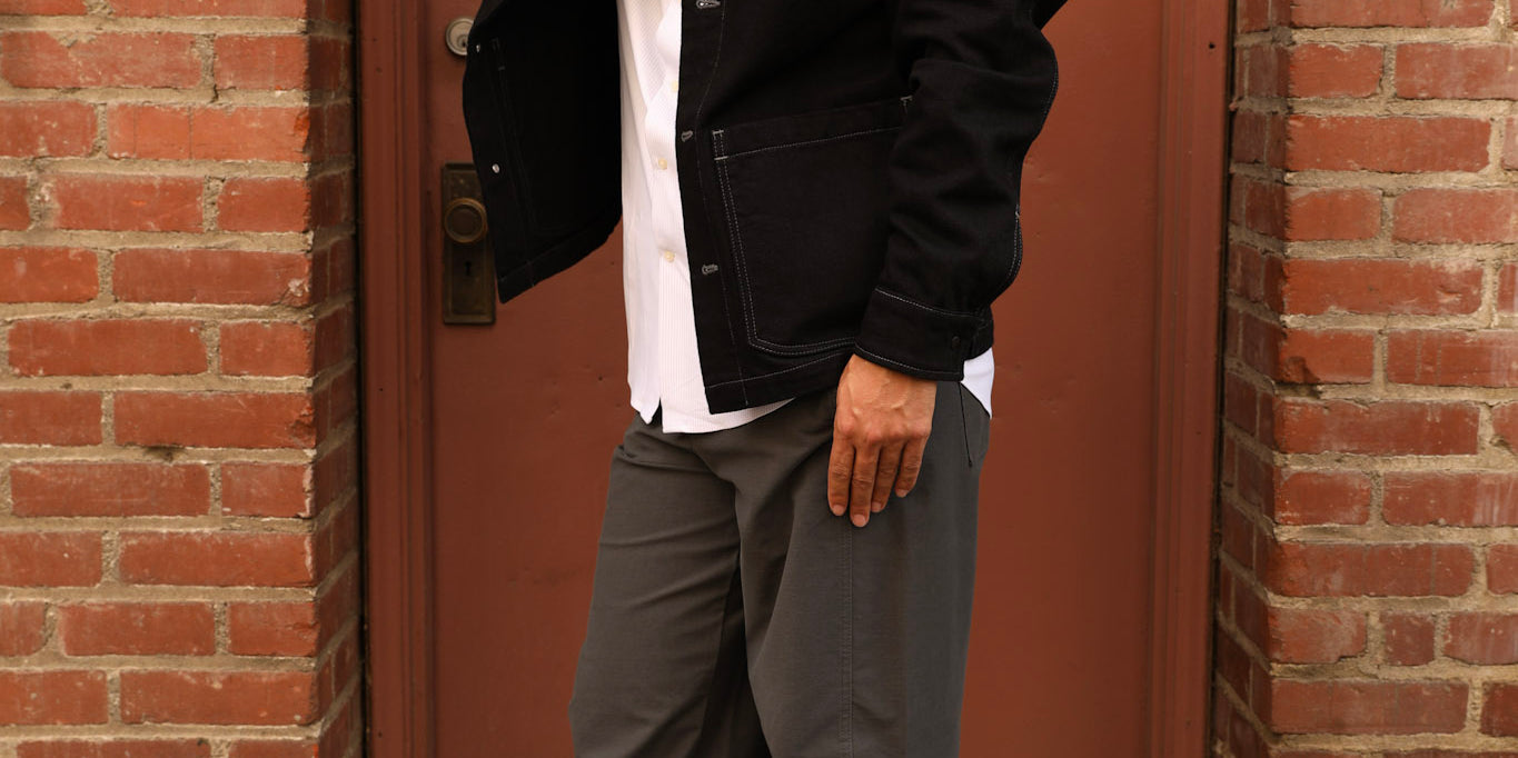 A man wearing a black baseball cap with a white 'A' logo, sunglasses, a black jacket over a white shirt, gray wide-leg pants, and black loafers, posing in front of a red brick door.