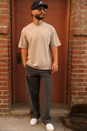 A man wearing a beige crewneck t-shirt, dark gray relaxed-fit pants, white sneakers, and a black baseball cap with a white 'A' logo, standing in front of a red-brown wooden door framed by brick walls.