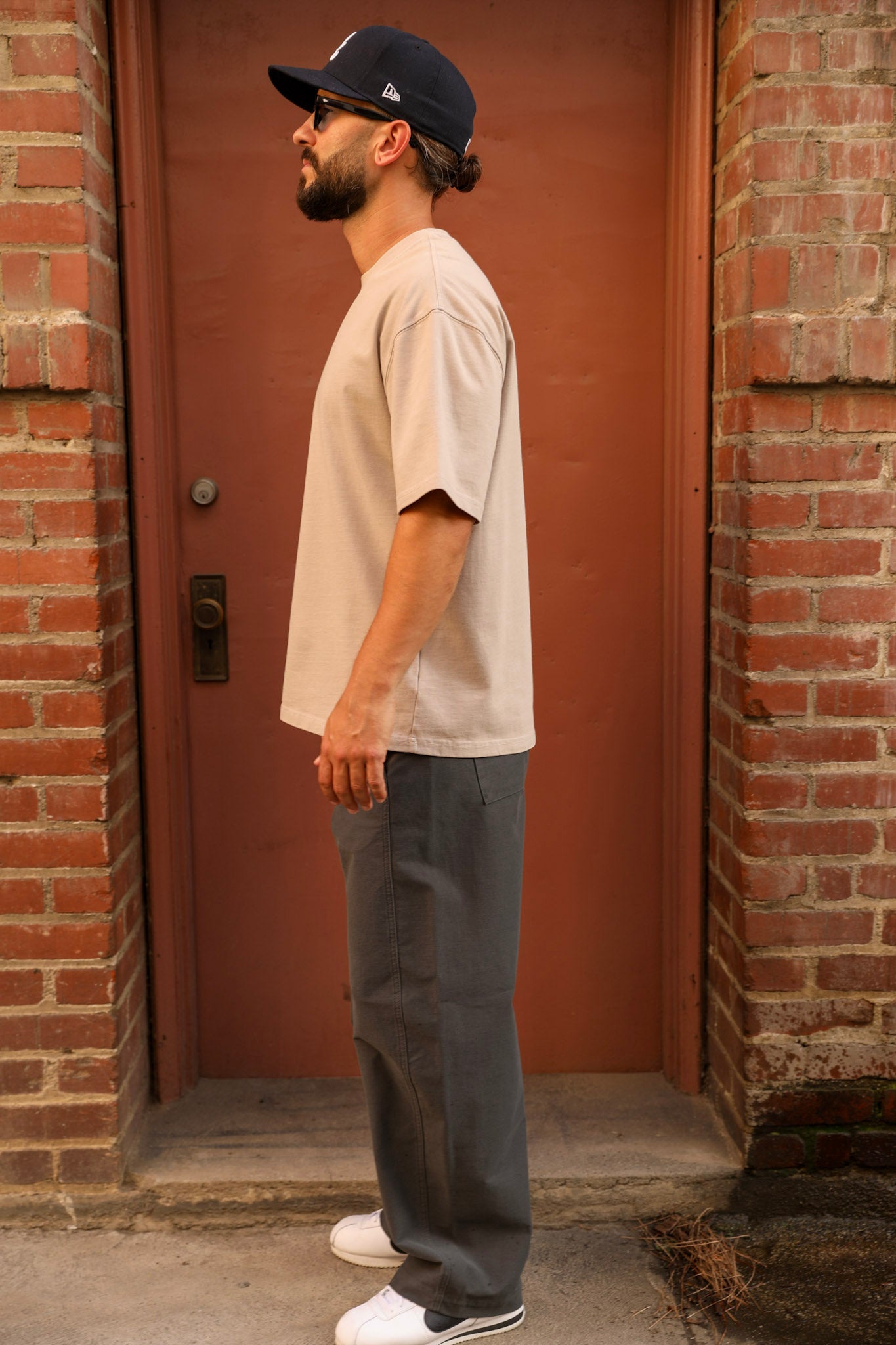 A man wearing a beige oversized t-shirt, dark gray relaxed-fit pants, white sneakers, and a black baseball cap stands in profile in front of a reddish-brown door framed by brick walls.