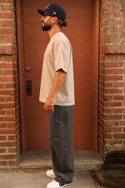 A man wearing a beige oversized t-shirt, dark gray relaxed-fit pants, white sneakers, and a black baseball cap stands in profile in front of a reddish-brown door framed by brick walls.