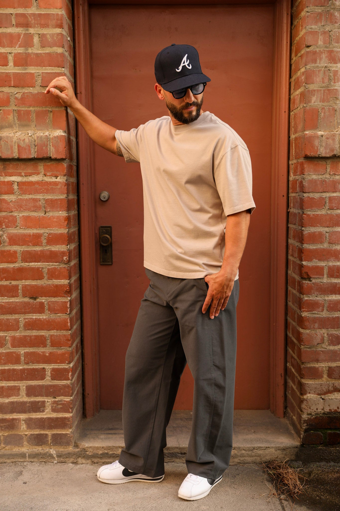A man wearing a beige t-shirt, dark gray wide-leg pants, a black baseball cap with a white 'A' logo, and white sneakers stands in front of a brick building with a rust-colored door.