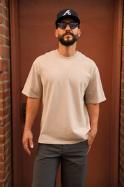 A man wearing a light beige crewneck t-shirt, dark gray pants, a black baseball cap with a white 'A' logo, and sunglasses, standing in front of a reddish-brown wooden door.
