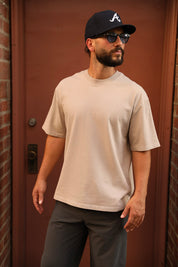 A man wearing a light beige crewneck t-shirt, dark gray pants, a black baseball cap with a white logo, and sunglasses, standing in front of a reddish-brown wooden door.