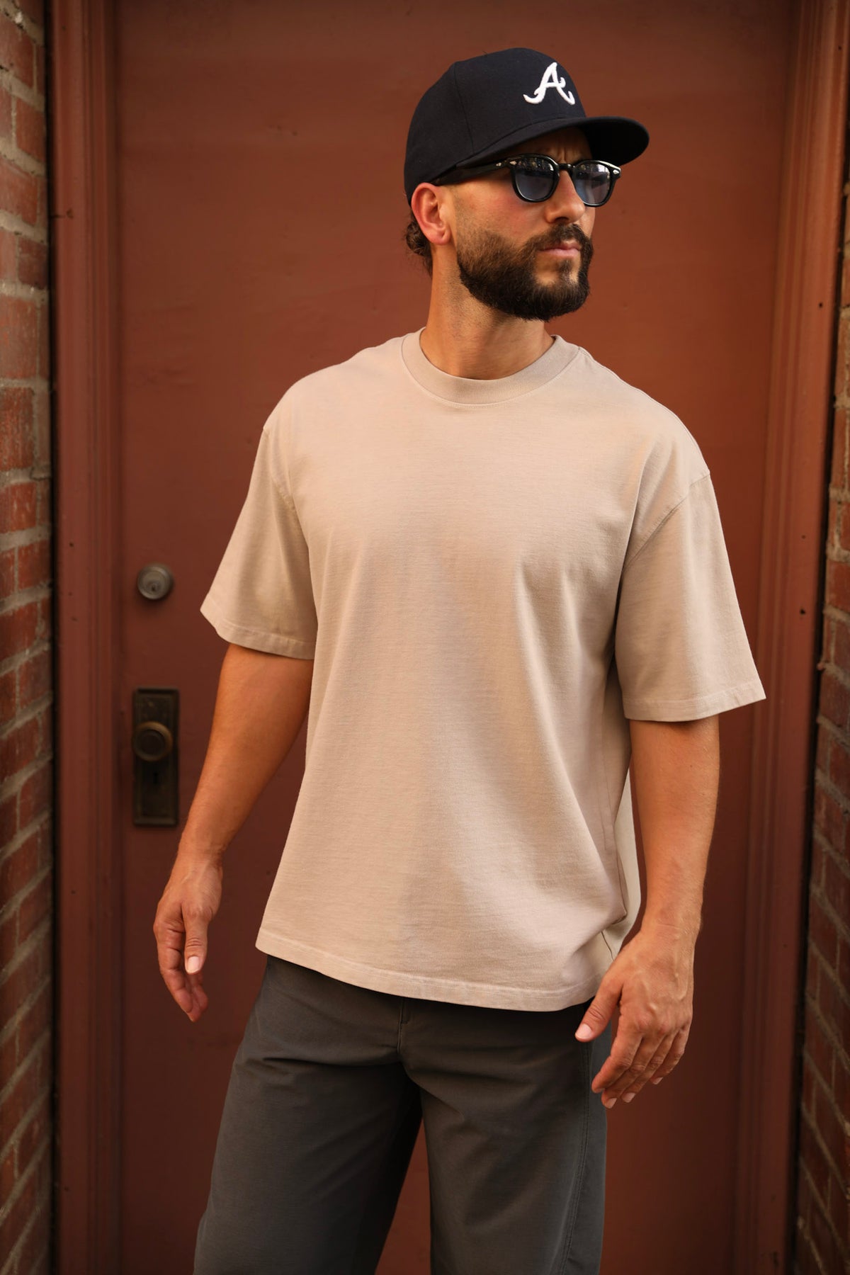 A man wearing a light beige crewneck t-shirt, dark gray pants, a black baseball cap with a white logo, and sunglasses, standing in front of a reddish-brown wooden door.