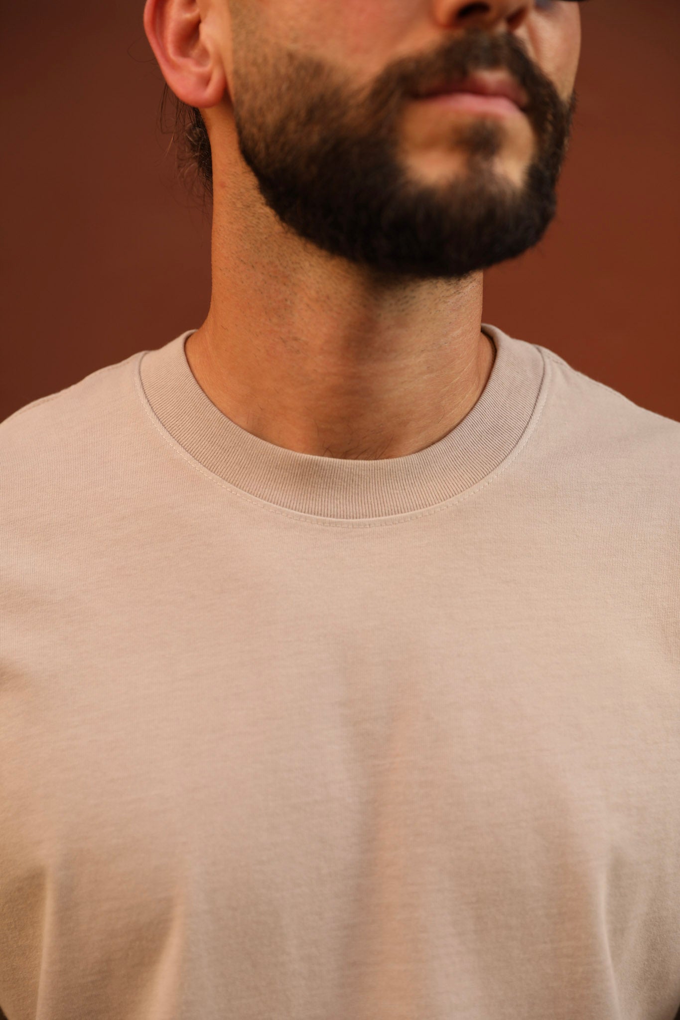 Close-up of a man wearing a light beige crew-neck t-shirt against a warm brown background.