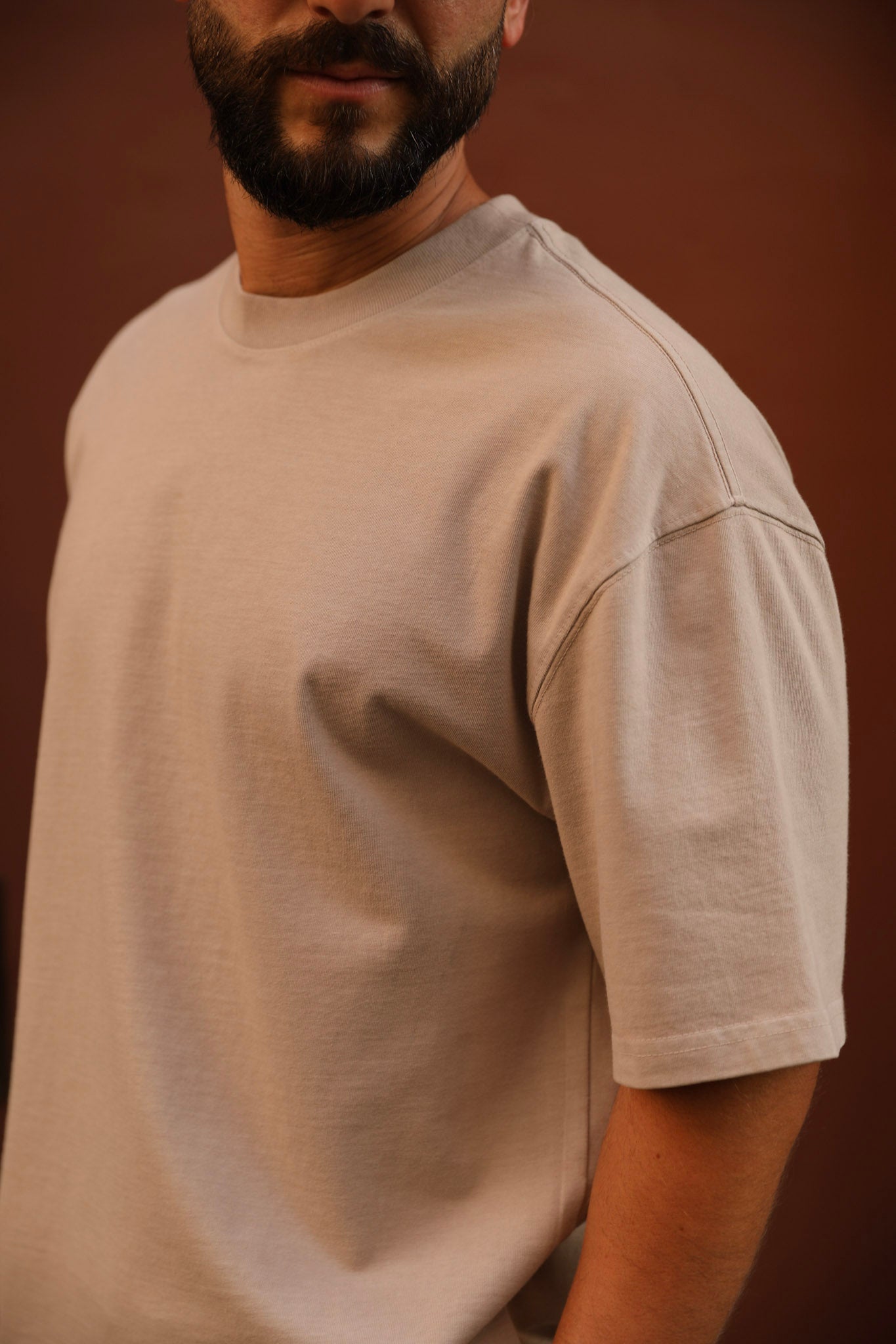 A man wearing a light beige crew-neck t-shirt, shown from the chest up against a solid dark brown background.