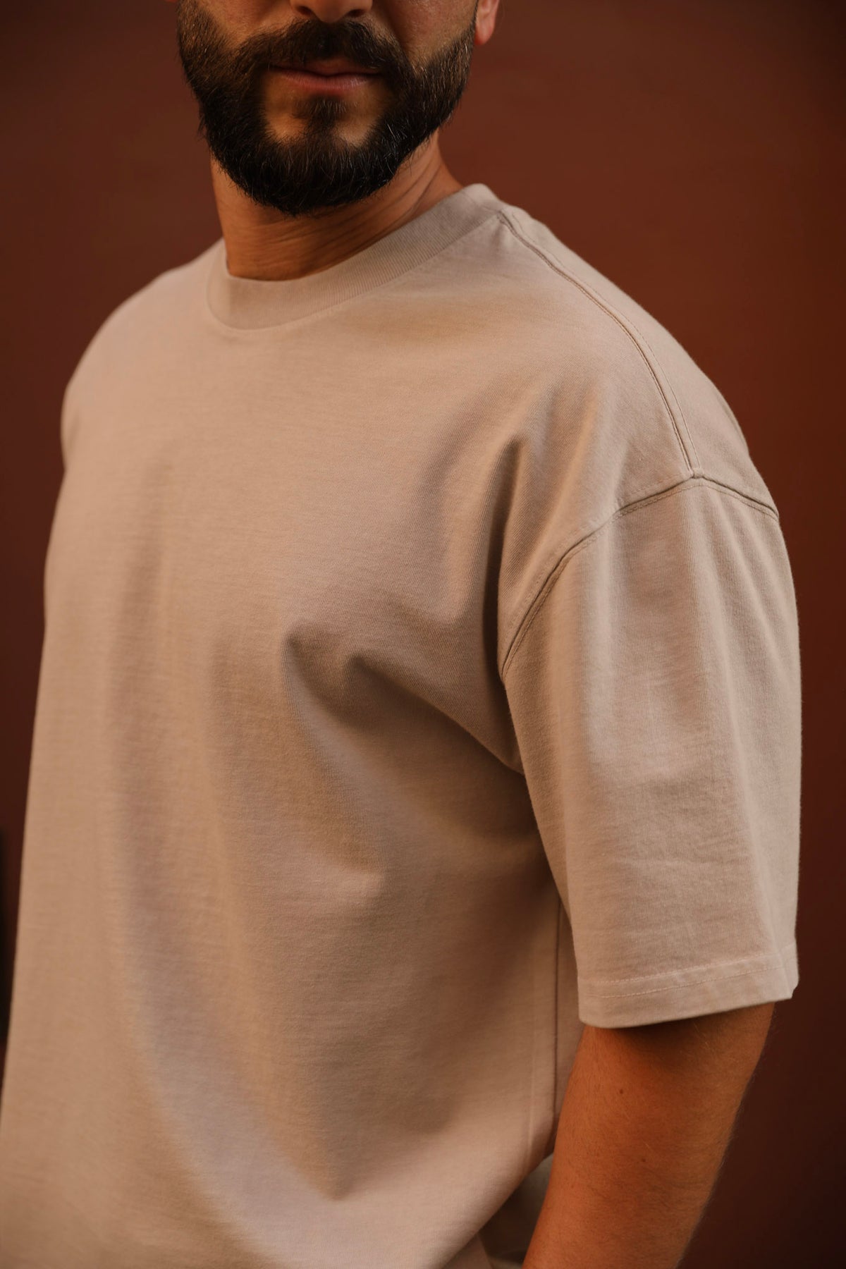 A man wearing a light beige crew-neck t-shirt, shown from the chest up against a solid dark brown background.