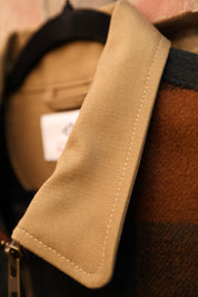 Close-up detail of a tan canvas collar with white contrast stitching, attached to a brown and black plaid wool jacket.