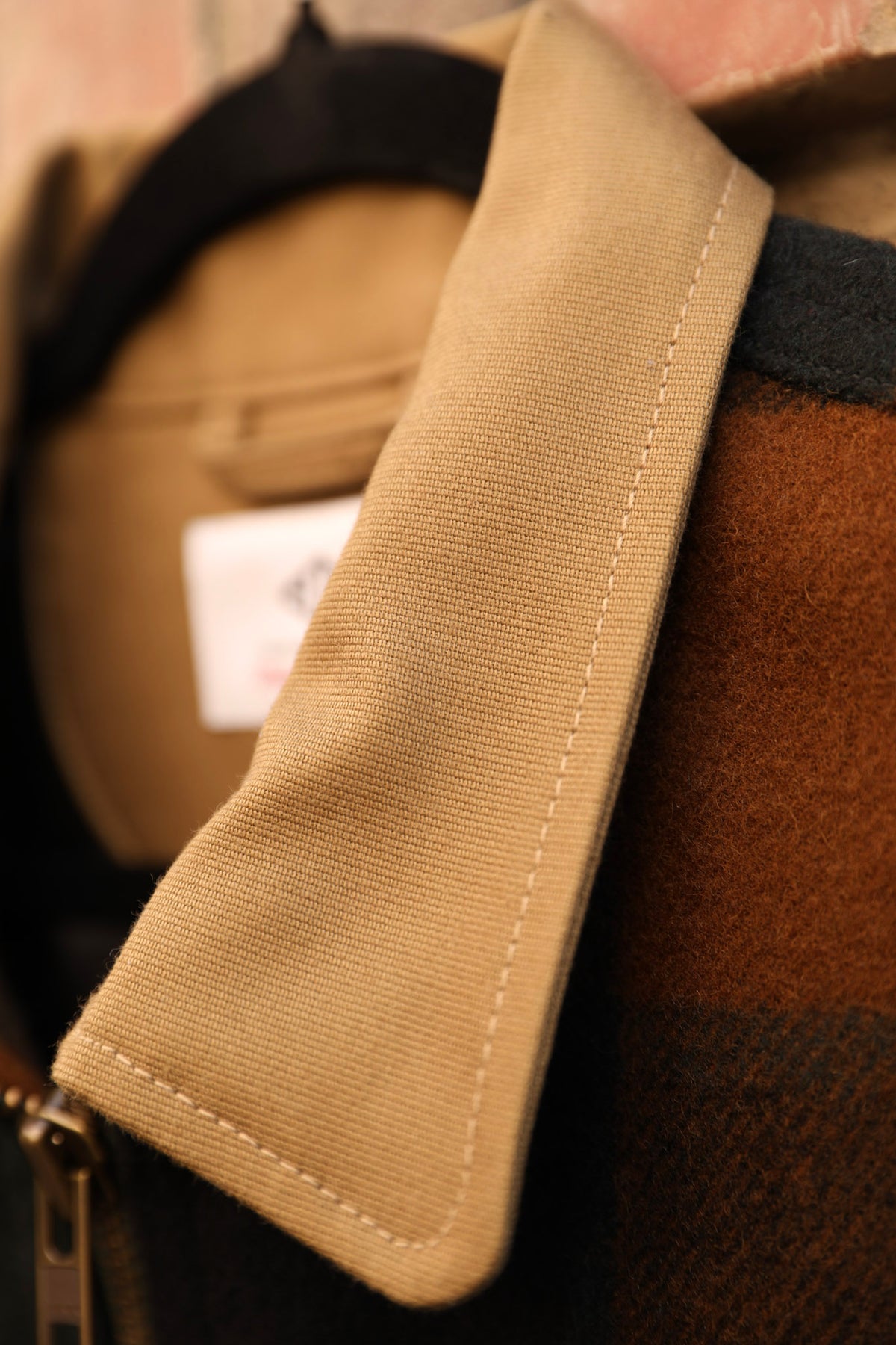 Close-up detail of a tan canvas collar with white contrast stitching, attached to a brown and black plaid wool jacket.