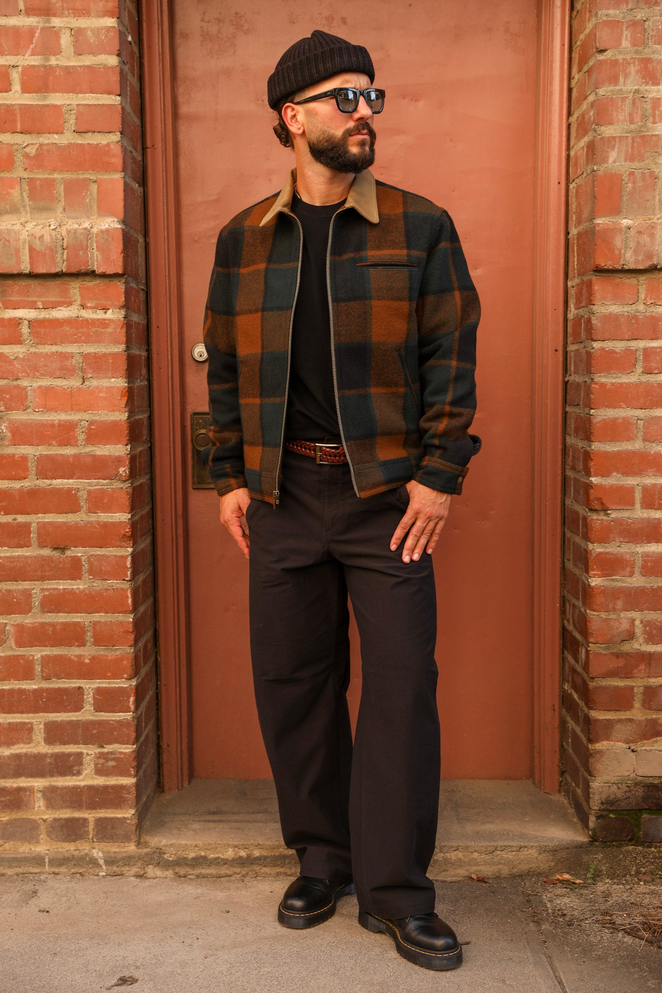 A man wearing a brown and black plaid overshirt, black pants, a black beanie, and sunglasses stands in front of a red-brown door framed by brick walls.