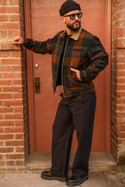 A man wearing a brown and black plaid zip-up jacket with a tan collar, dark trousers, a black beanie, and sunglasses, posing in front of a brick wall and reddish-brown door.