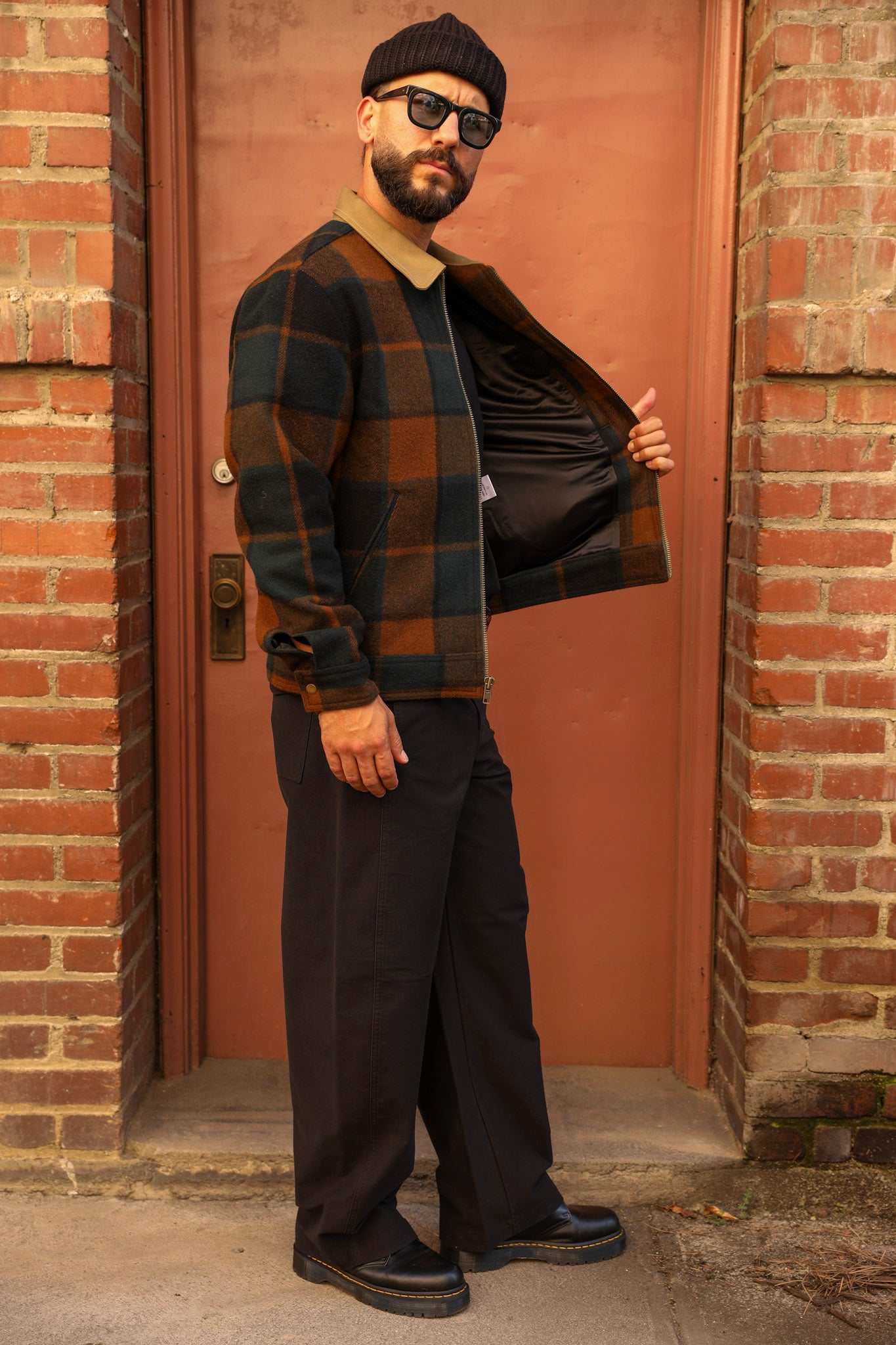 A man wearing a black beanie, sunglasses, a brown and black plaid overshirt over a tan shirt, and dark wide-leg trousers stands in front of a reddish-brown door framed by brick walls.