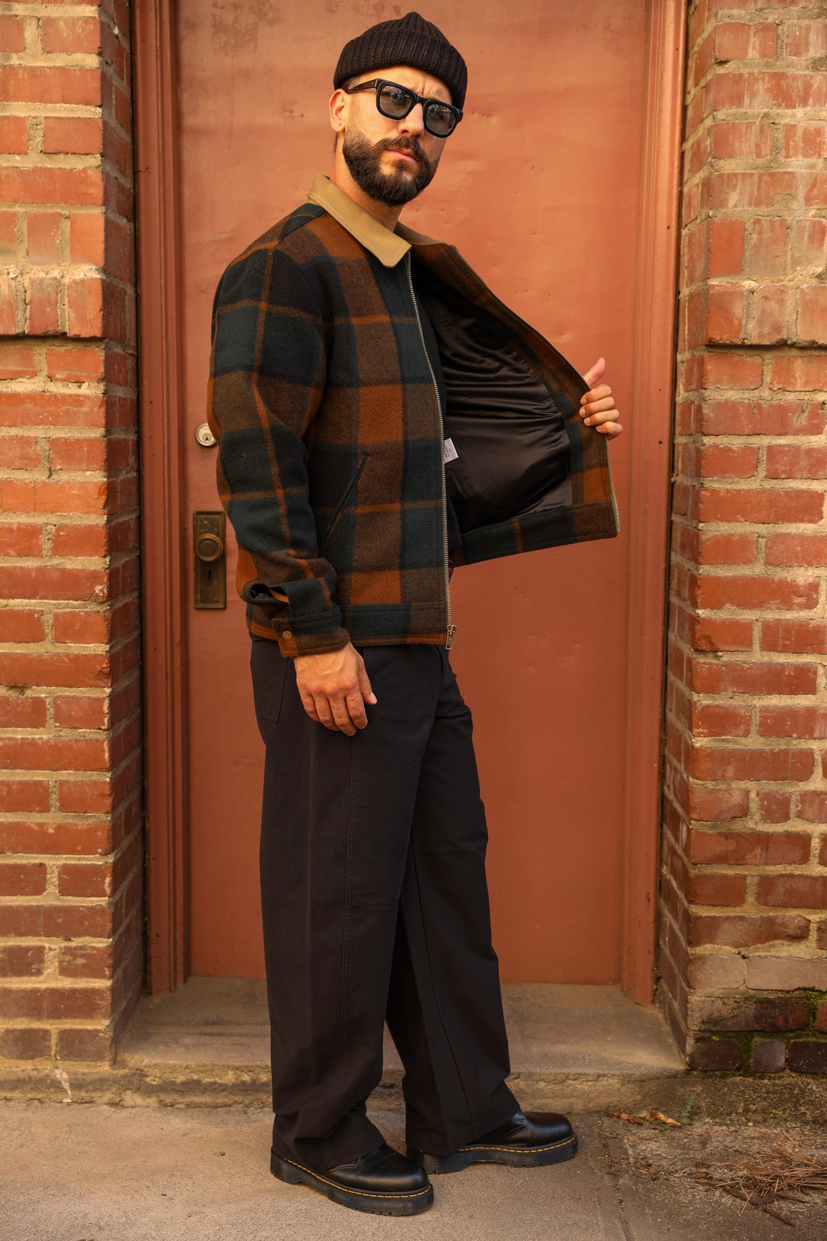A man wearing a black beanie, sunglasses, a brown and black plaid overshirt over a tan shirt, and dark wide-leg trousers stands in front of a reddish-brown door framed by brick walls.