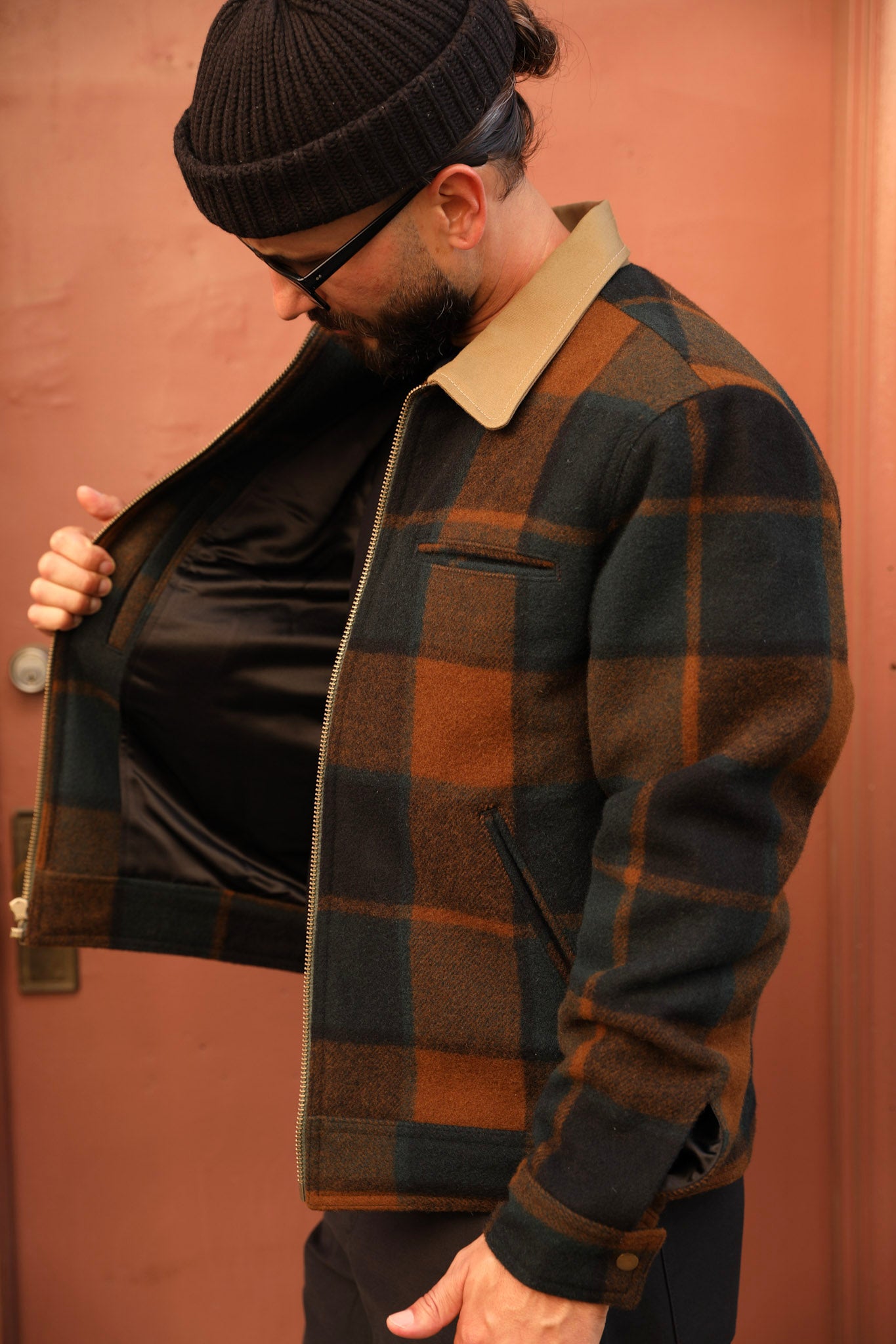 A man wearing a dark brown and black plaid wool jacket with a tan collar, shown opening the jacket to reveal a black satin lining, set against a warm terracotta-colored wall.