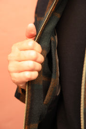 Close-up of a person's hand holding open a dark green and brown plaid zippered jacket over a black top, against a solid peach-colored background.