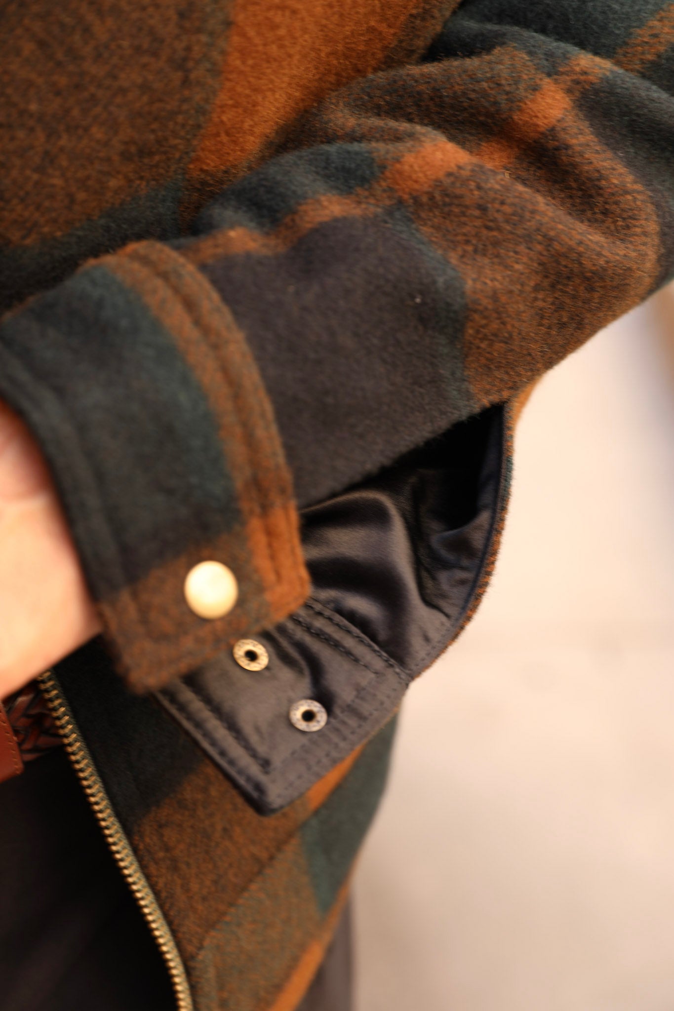 Close-up of a person wearing a dark green and brown plaid flannel jacket, showing the sleeve cuff with snap buttons and a black interior lining.