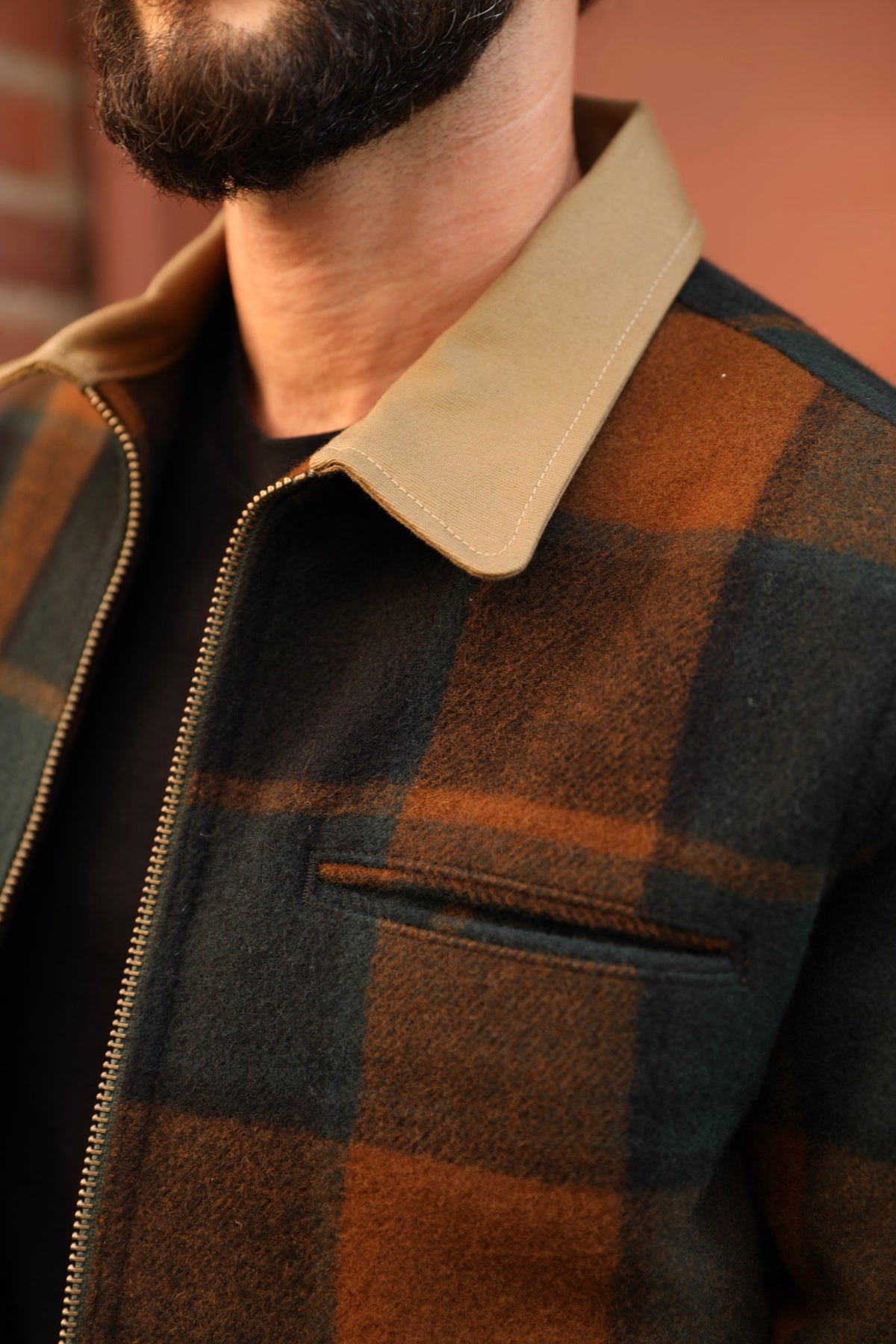 Close-up of a man wearing a brown and black buffalo plaid zip-up jacket with a tan corduroy-style collar over a black shirt.