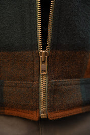 Close-up of a vintage-style zippered jacket featuring a dark gray wool body, a brown and orange buffalo plaid pattern, and a brass-colored metal zipper.